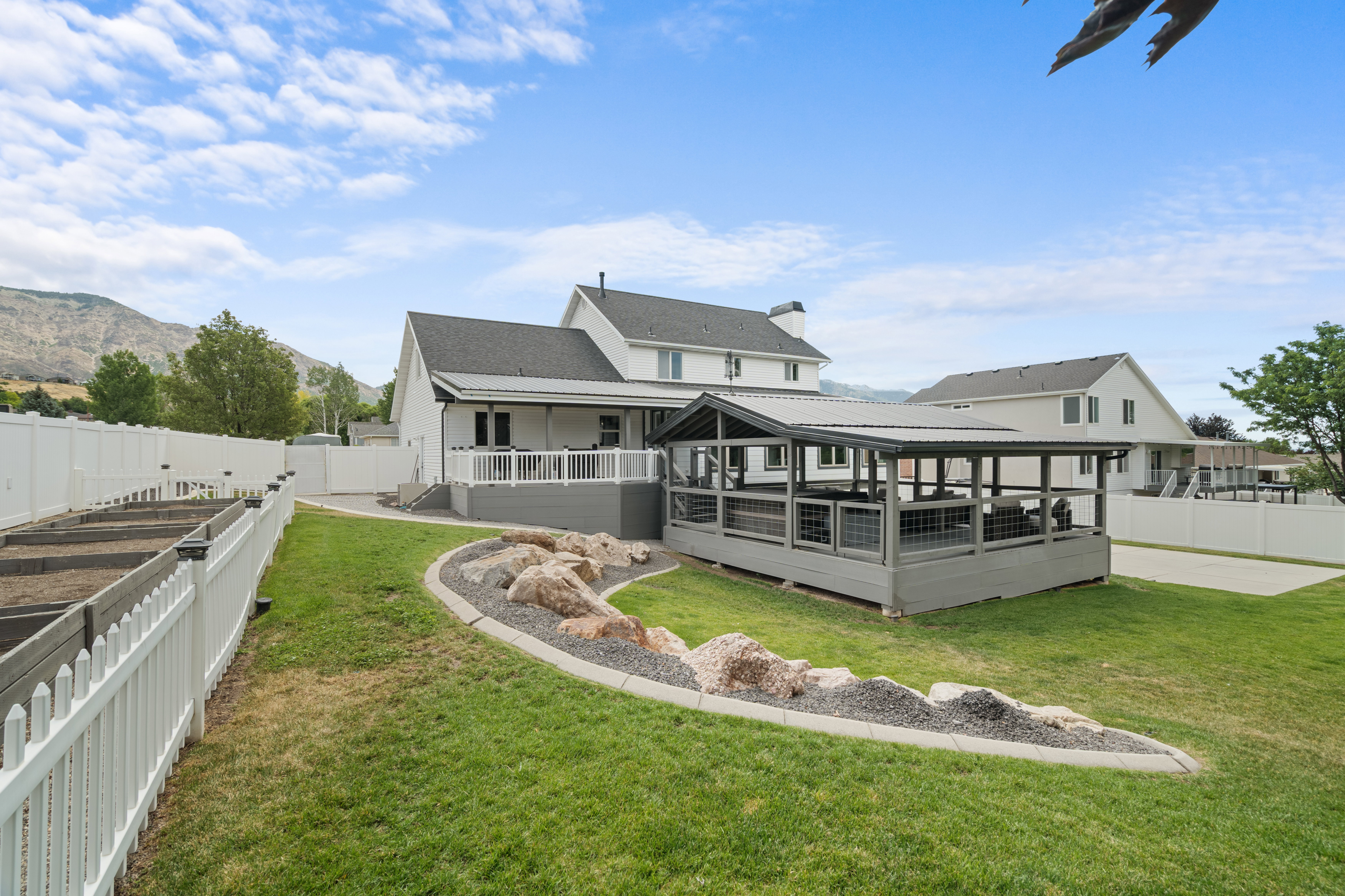 Spacious backyard featuring a covered patio, elevated deck with white railings, and a beautifully landscaped rock garden. The yard also includes eight fenced raised garden beds, providing ample space for gardening, flowers, or homegrown produce