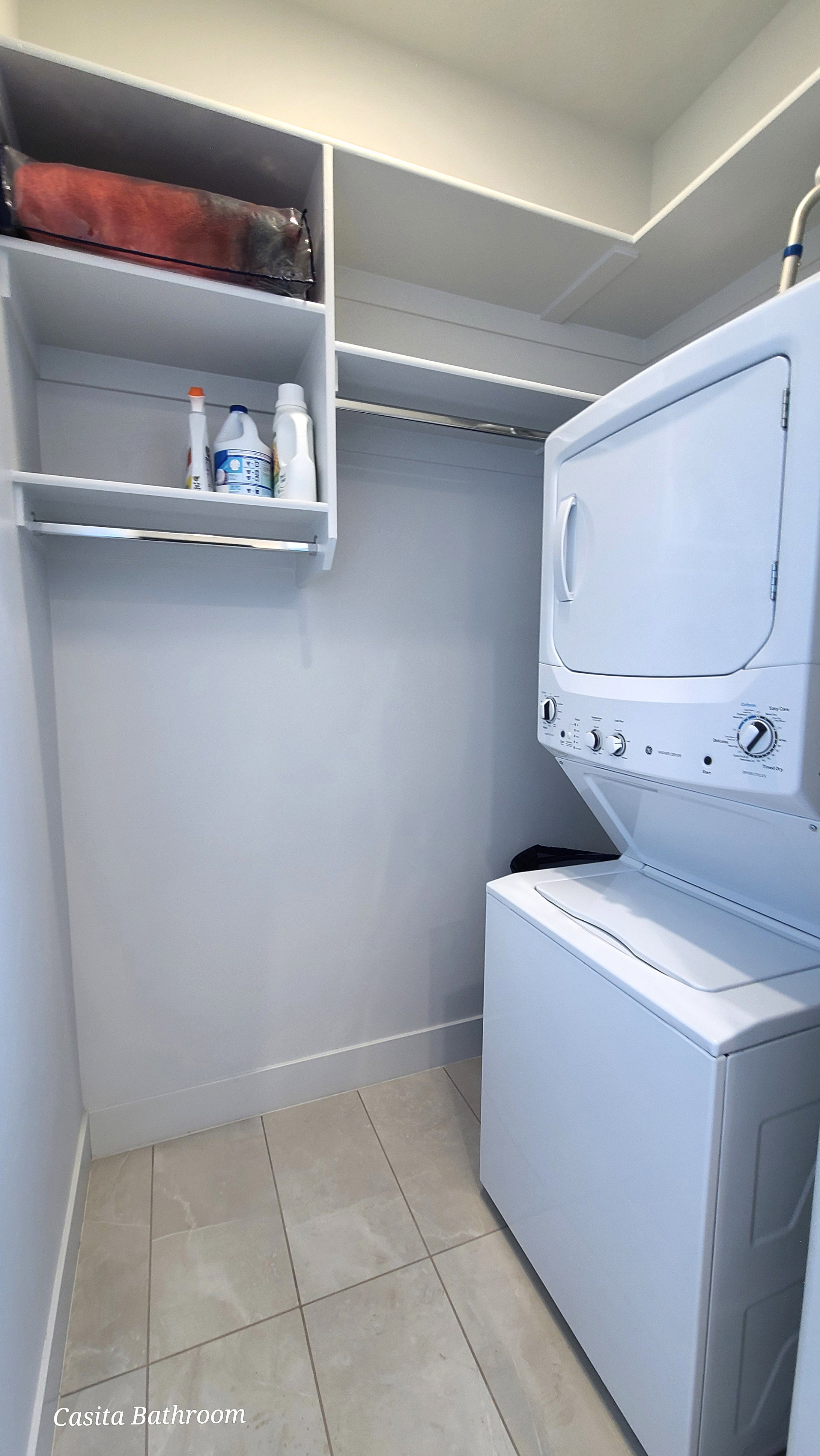 Dedicated laundry area featuring a stacked washer and dryer, built-in shelving, and light-toned floor tiling