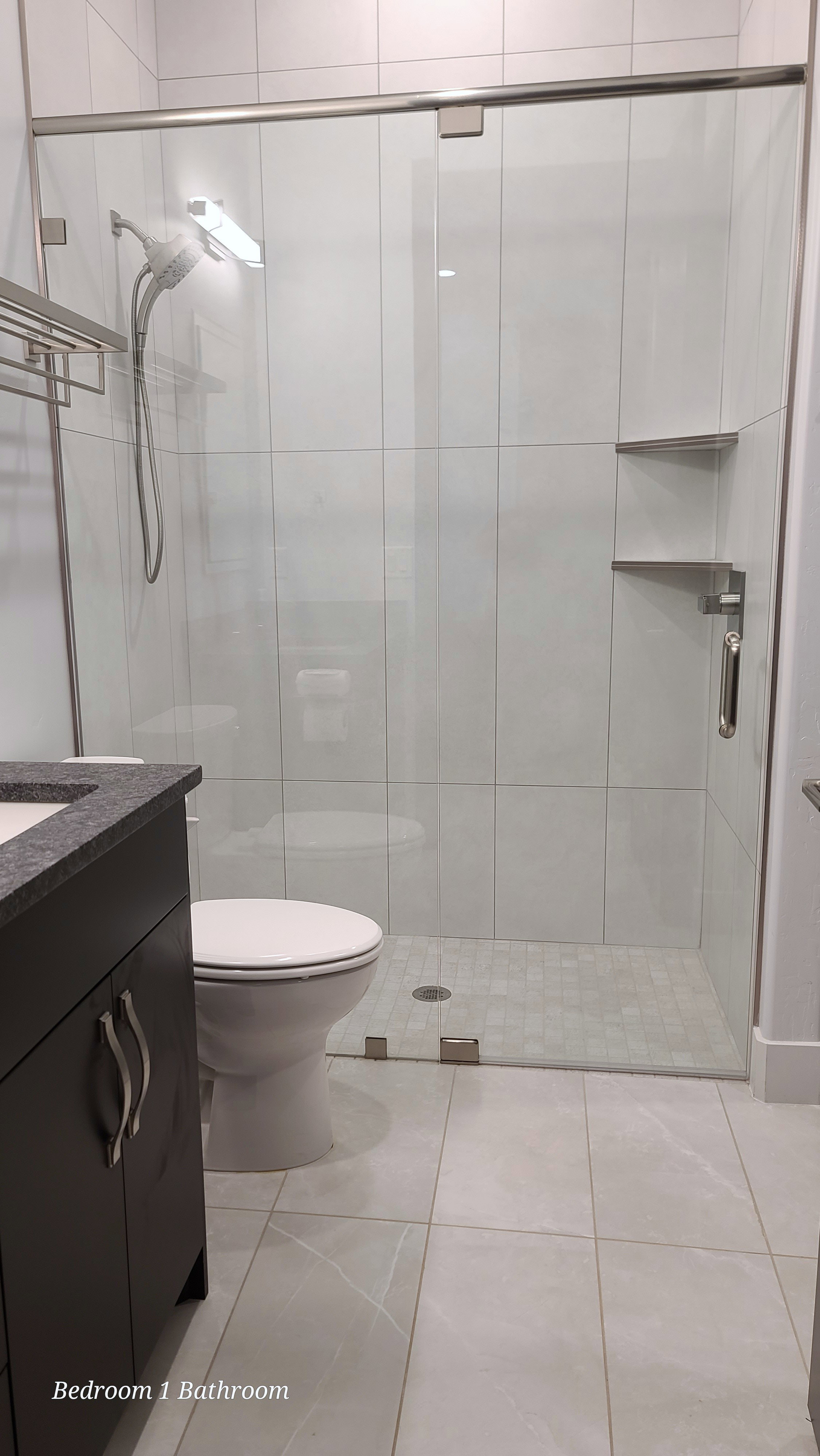Glass-enclosed shower with white tiled walls and built-in shelving, complemented by a dark vanity with a stone countertop and tile flooring