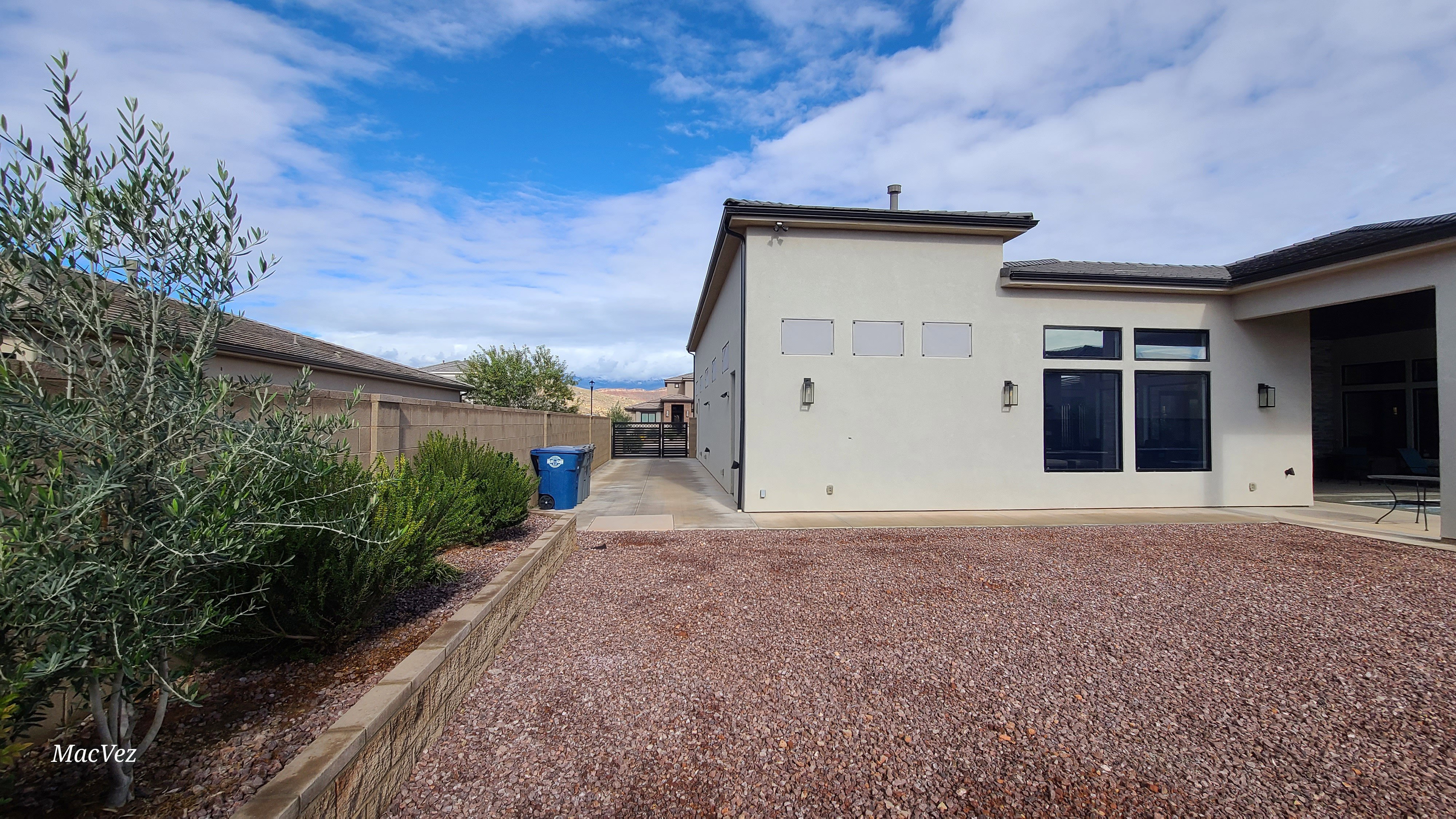 Spacious backyard featuring a low-maintenance gravel ground cover, a large stucco exterior, black-framed windows, exterior lighting fixtures, and a side access gate