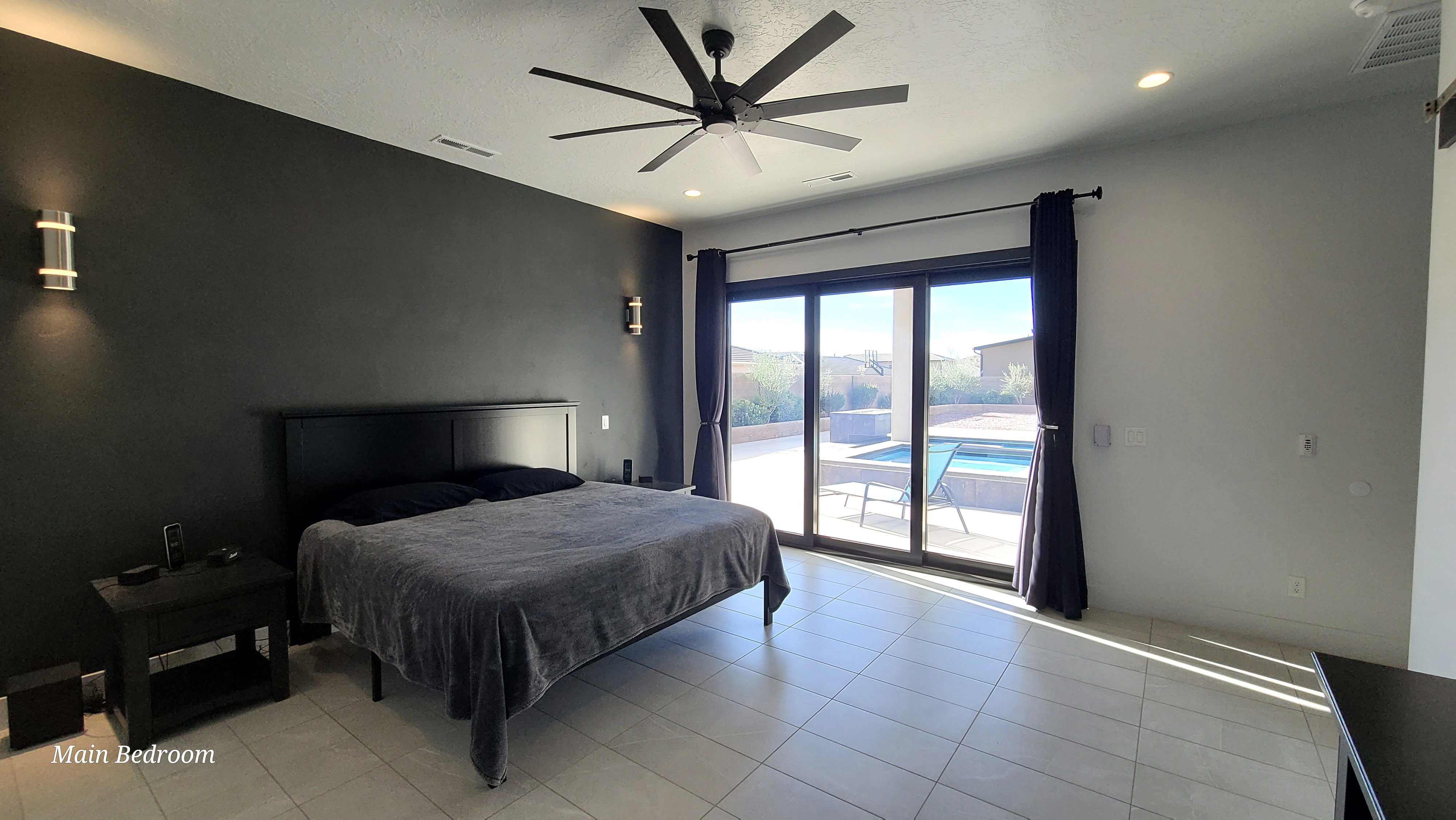 Spacious room featuring a dark accent wall with sconce lighting, an overhead ceiling fan, tile flooring, and large sliding glass doors opening to an outdoor pool and patio area