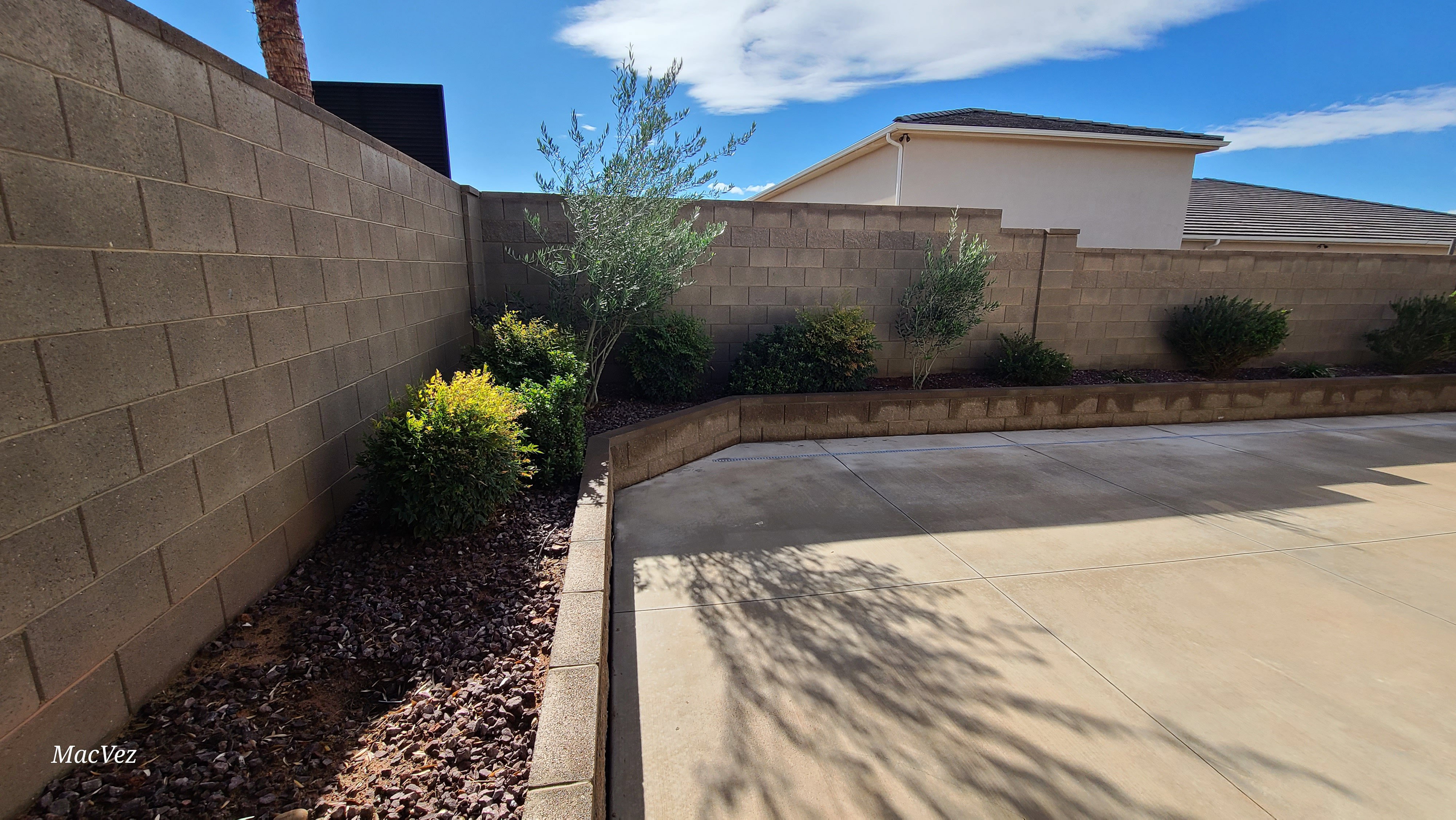 Paved outdoor area featuring concrete block walls and a rock-mulched planter bed with established shrubs and a young tree