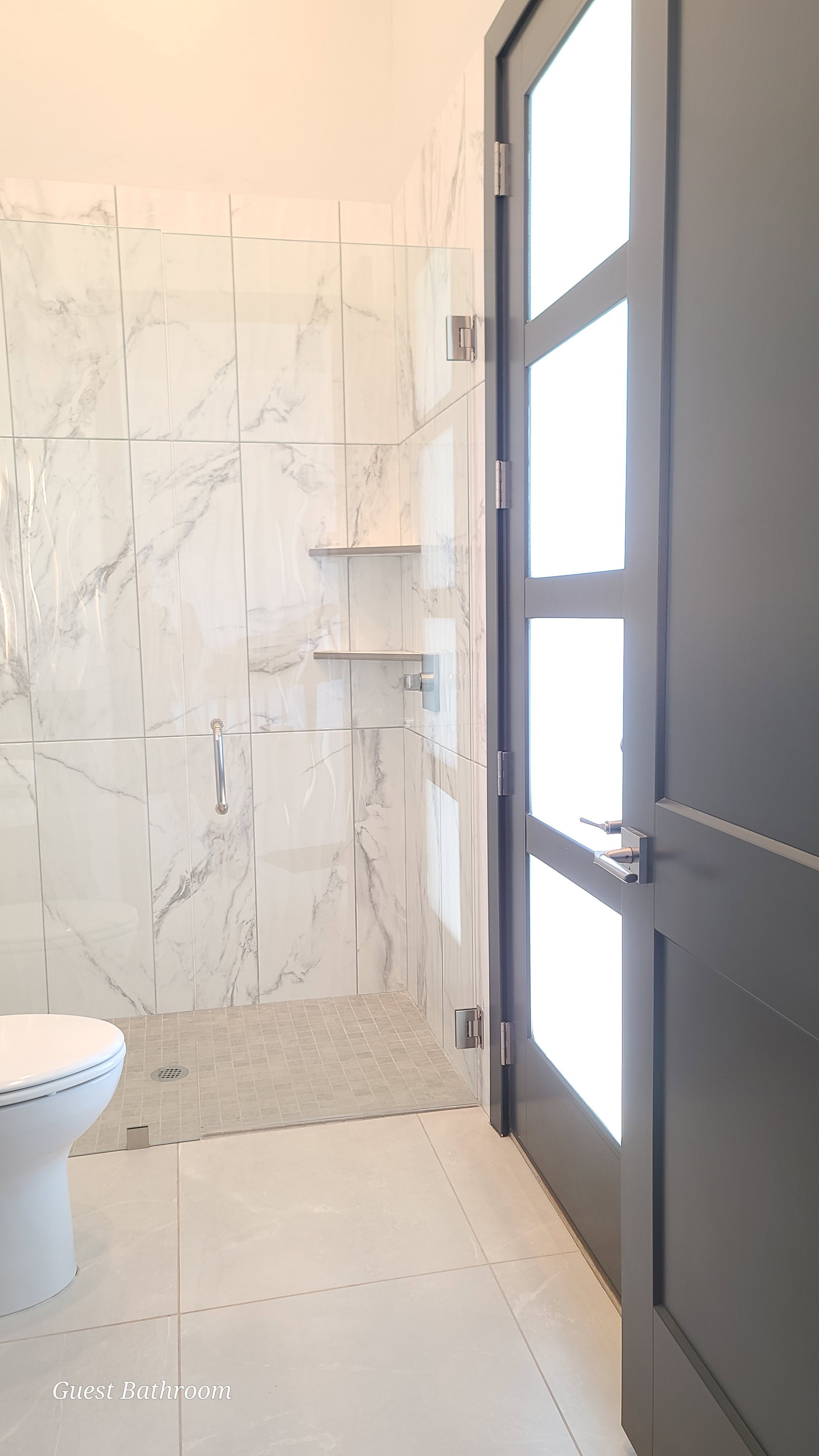 Spacious bathroom featuring a glass-enclosed shower with white marble-look wall tiles, built-in shelving, and a mosaic tile floor