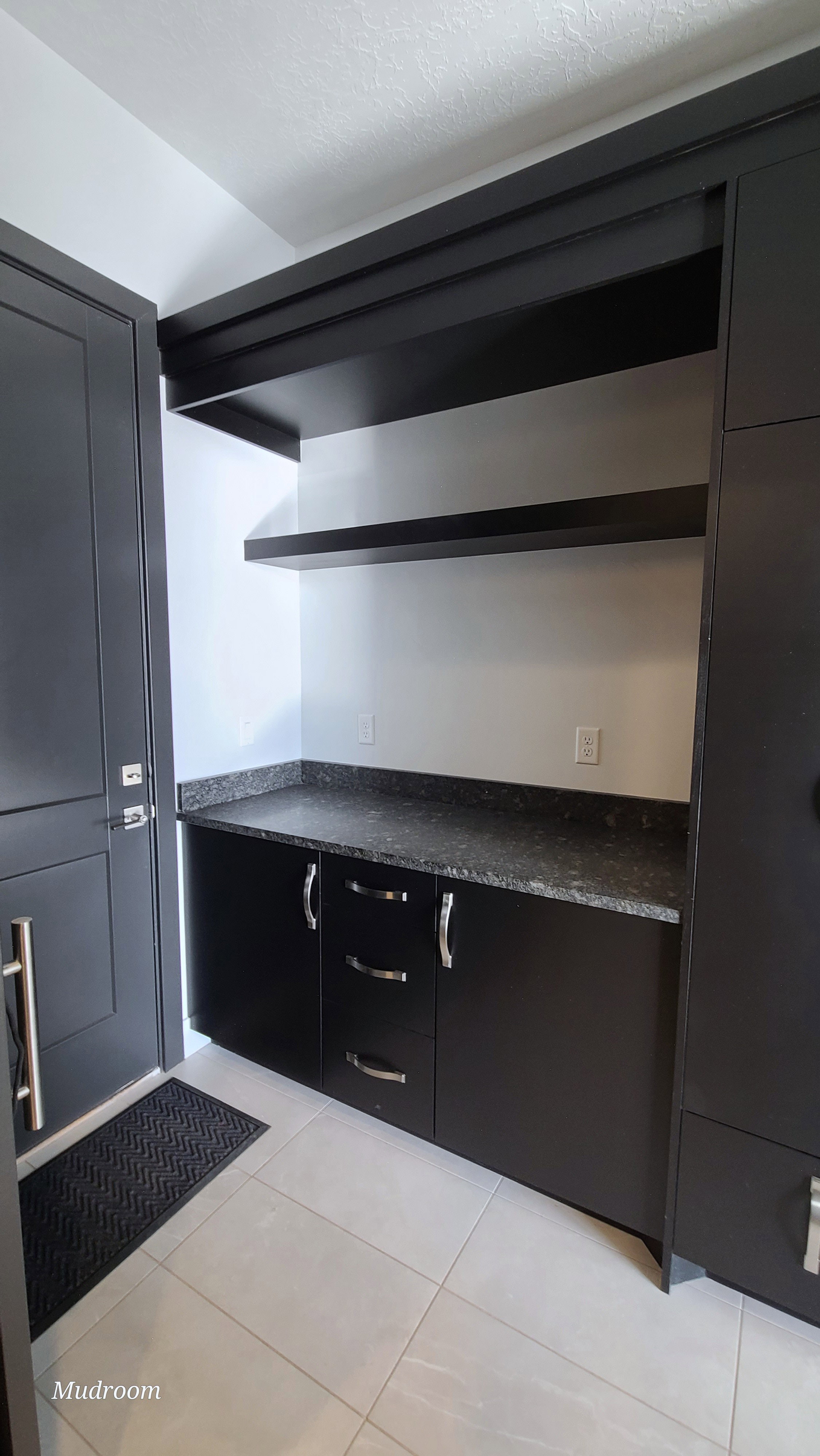 Entryway featuring dark cabinetry, granite-style countertops, floating shelves, and light tile flooring