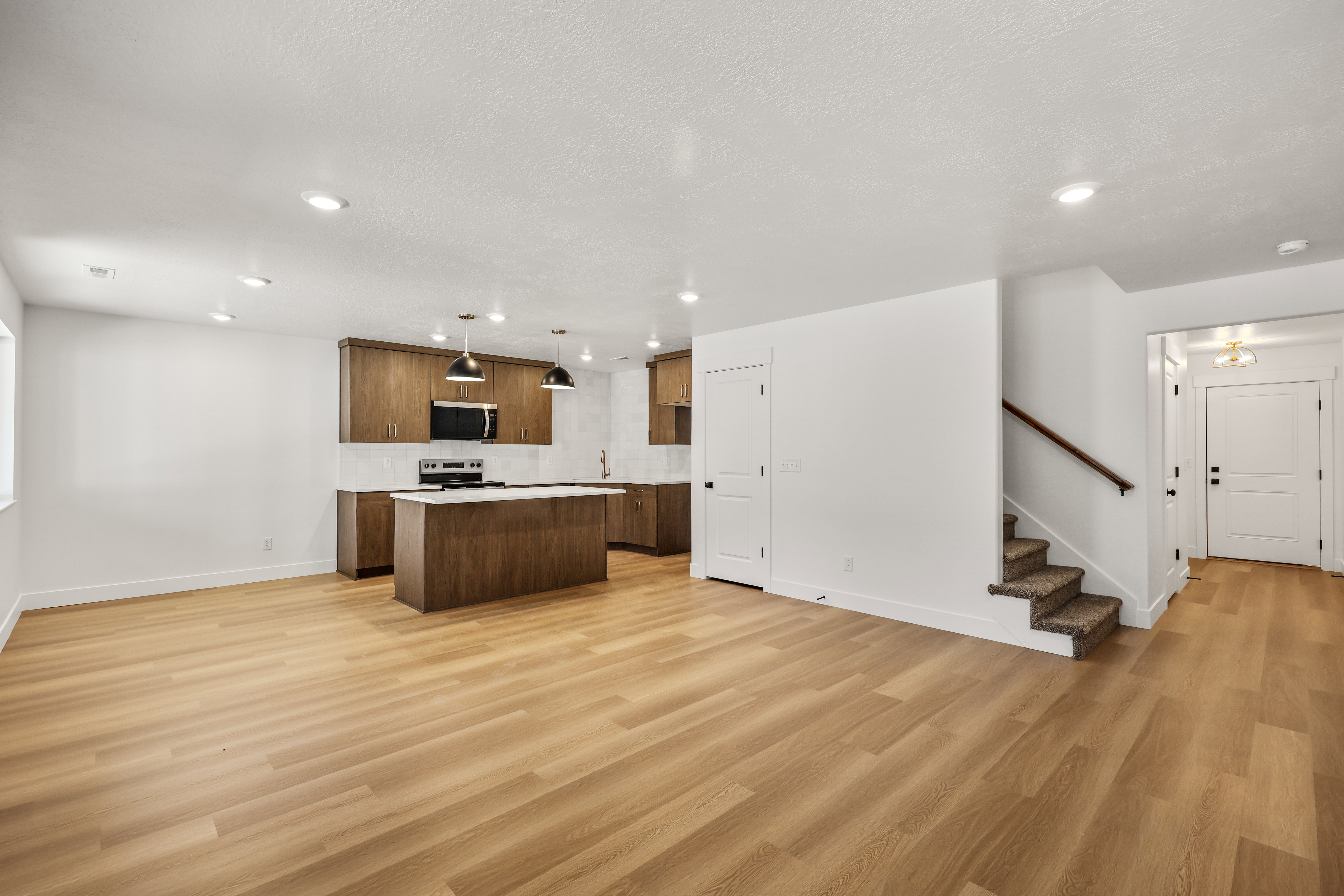 Open concept living space featuring wood-finish flooring, a kitchen with dark wood cabinetry and white countertops, stainless steel appliances, recessed ceiling lighting, and a carpeted staircase with a wood handrail