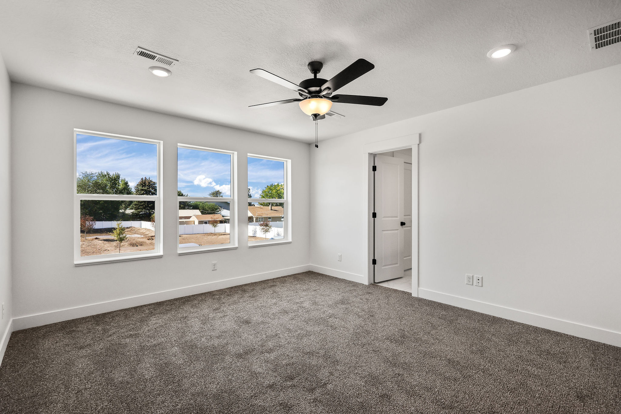 Room with gray carpeting and white walls, featuring three large windows, a ceiling fan with light fixture, and recessed lighting