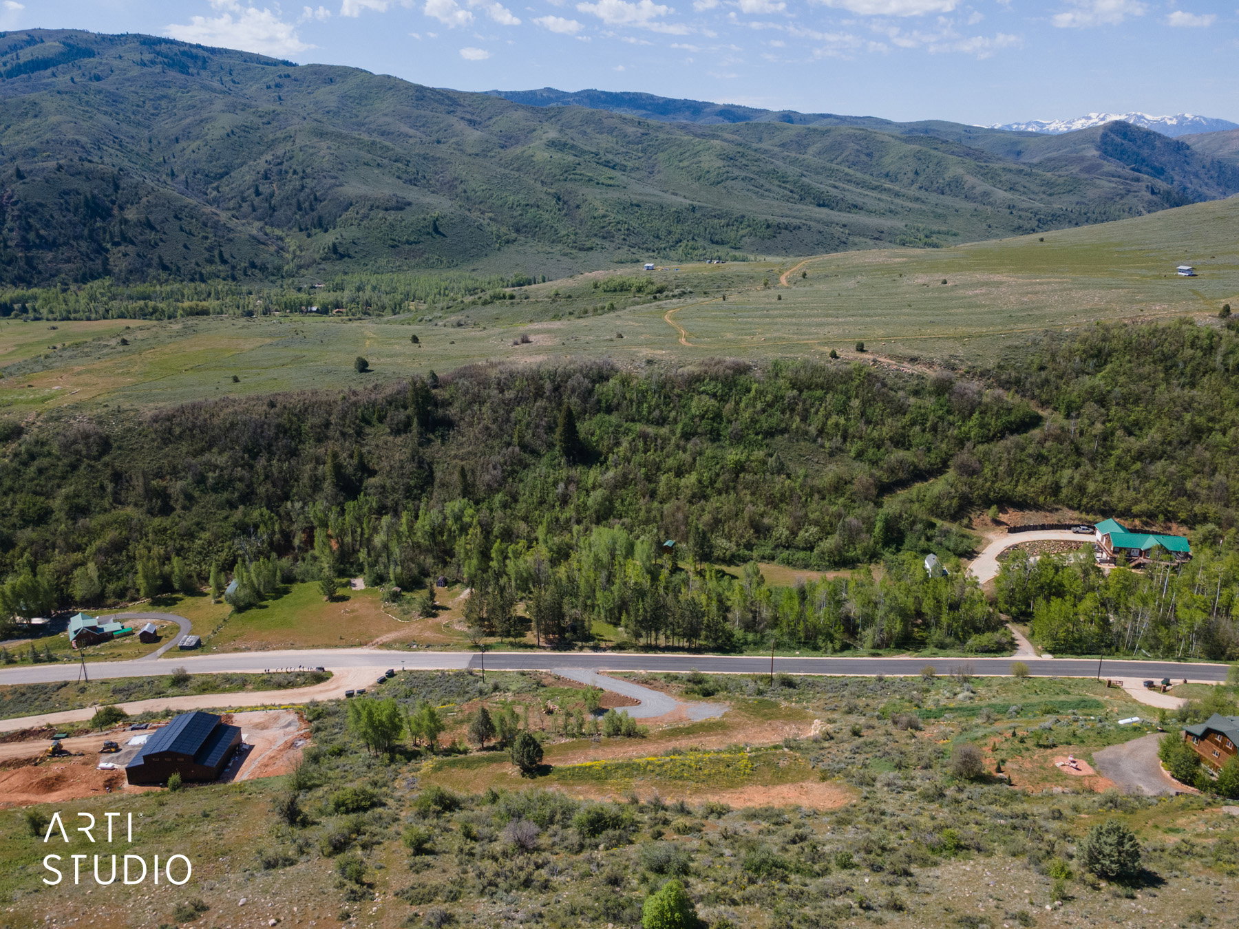 Expansive mountain property featuring varied terrain, mature trees, open fields, a paved road, and a distant snow-capped mountain range