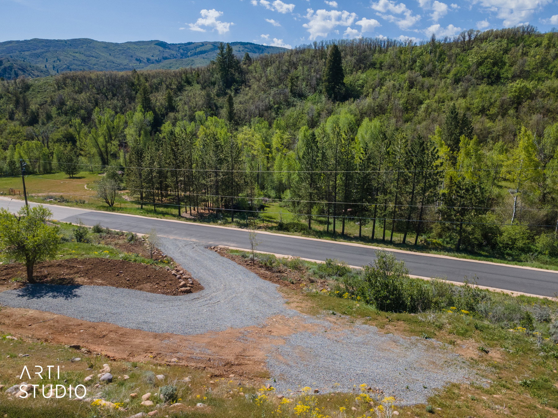 Gravel driveway with a paved road frontage, bordered by abundant mature trees and distant mountains