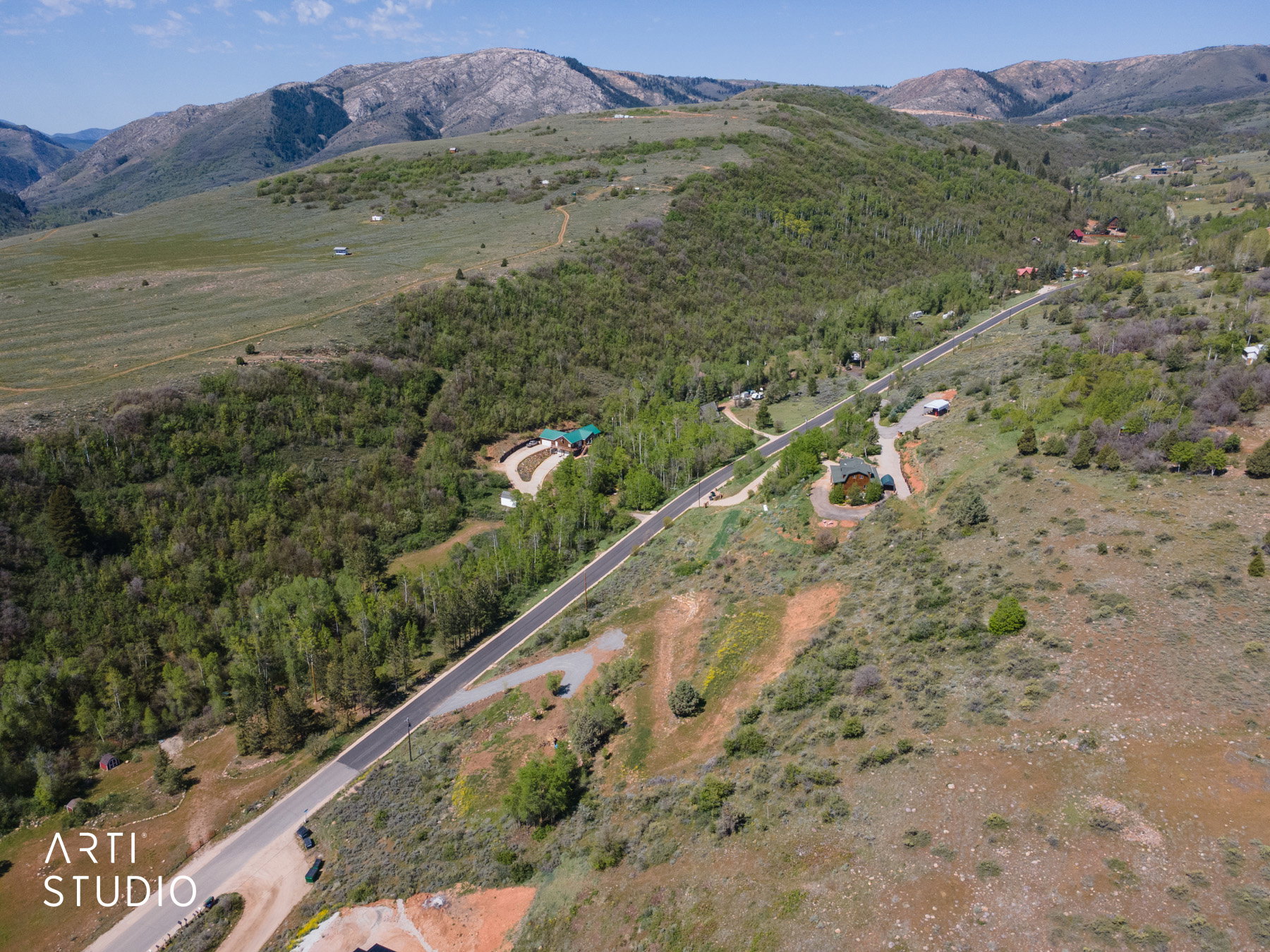 Expansive mountain landscape featuring a paved road traversing rolling hills, abundant mature trees, and distant mountain ranges