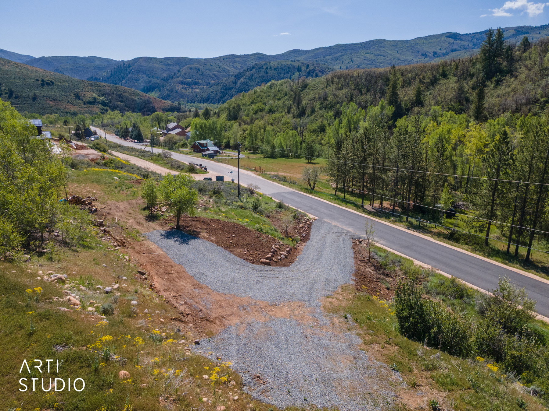 Gravel driveway entrance with a paved road running alongside, surrounded by green hillsides and mature trees