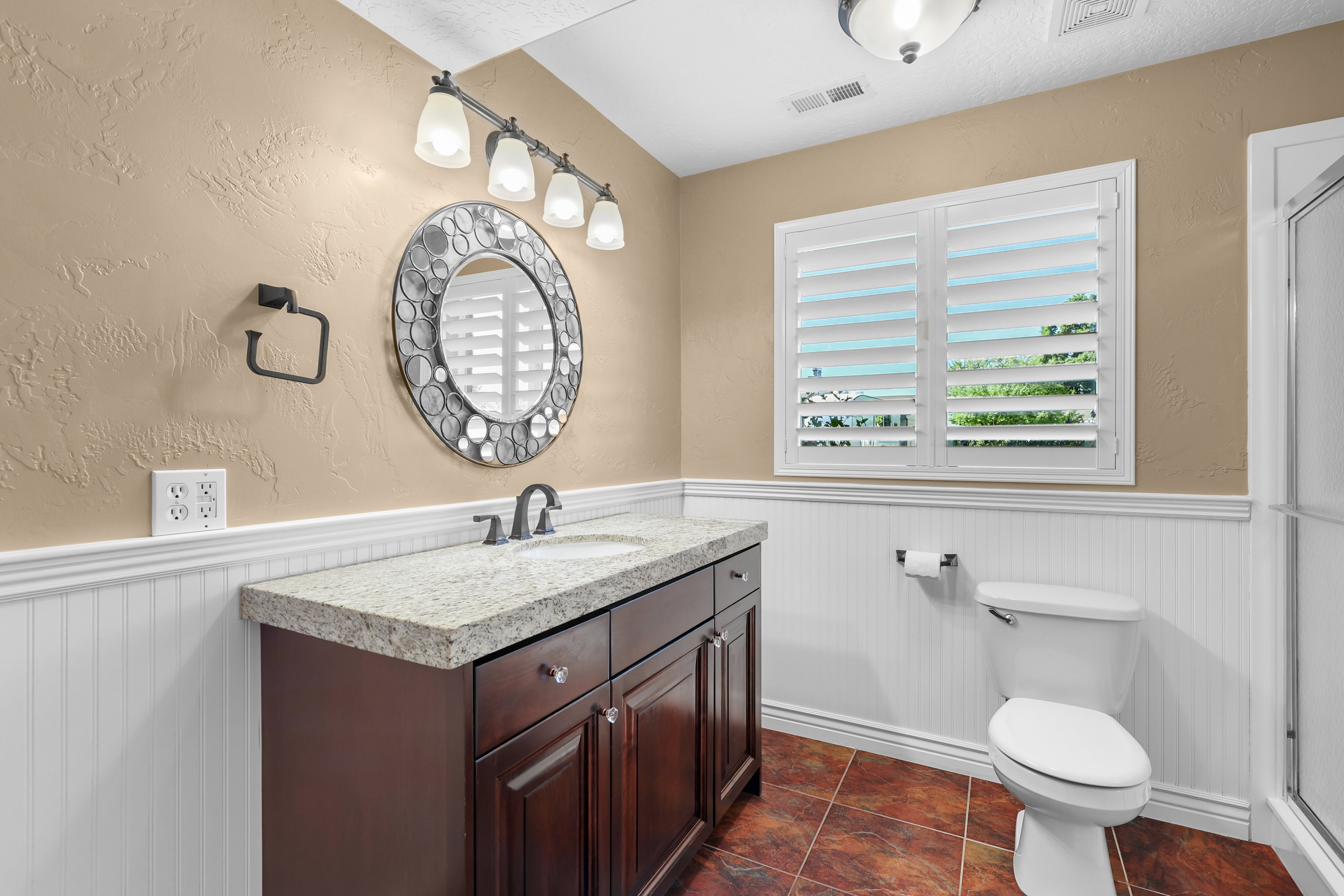 Bathroom featuring a dark wood vanity with a light granite countertop, a decorative round mirror, and a window with white plantation shutters