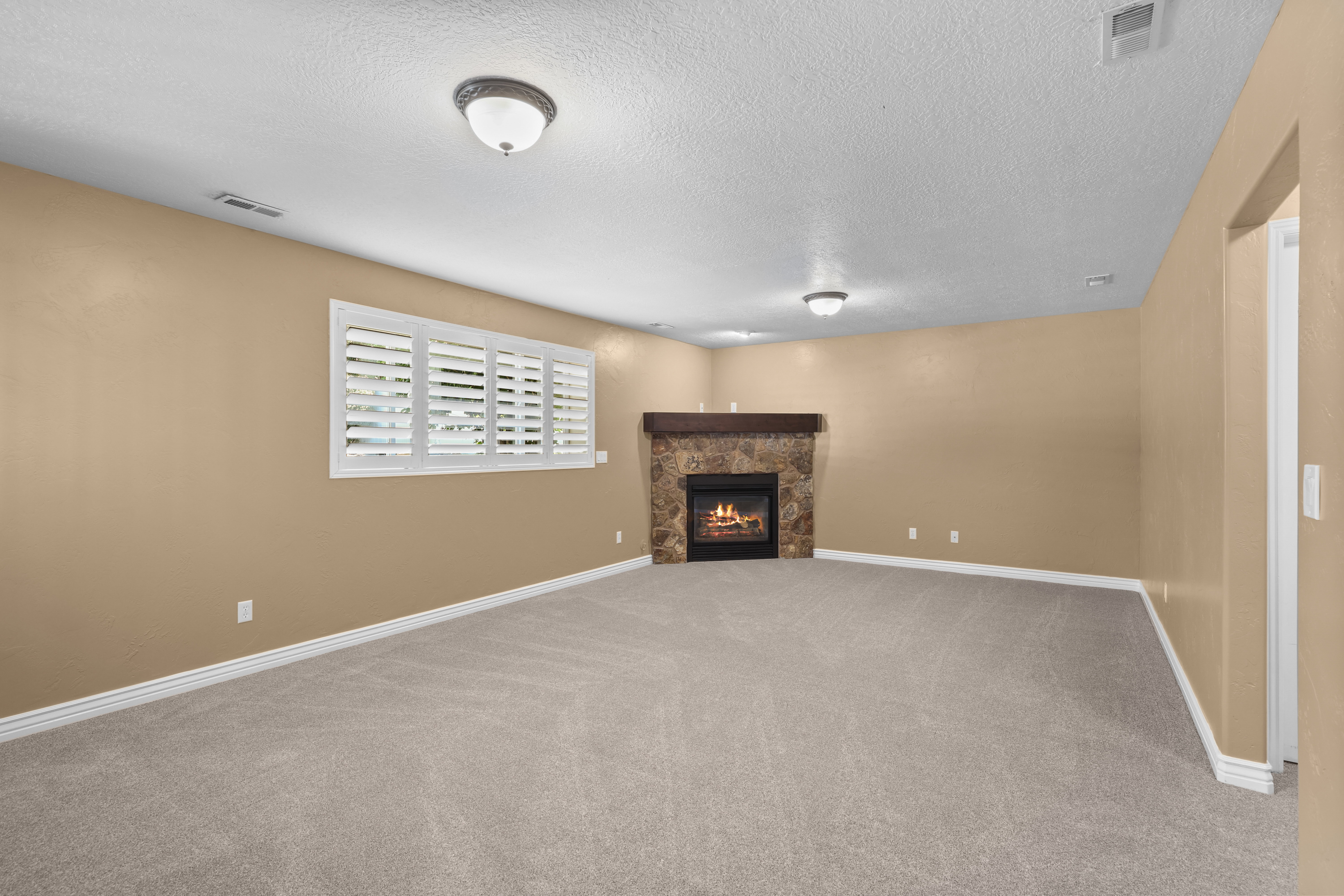 Spacious interior room featuring a stone fireplace with a wood-finish mantel, neutral carpeting, and a window with plantation shutters