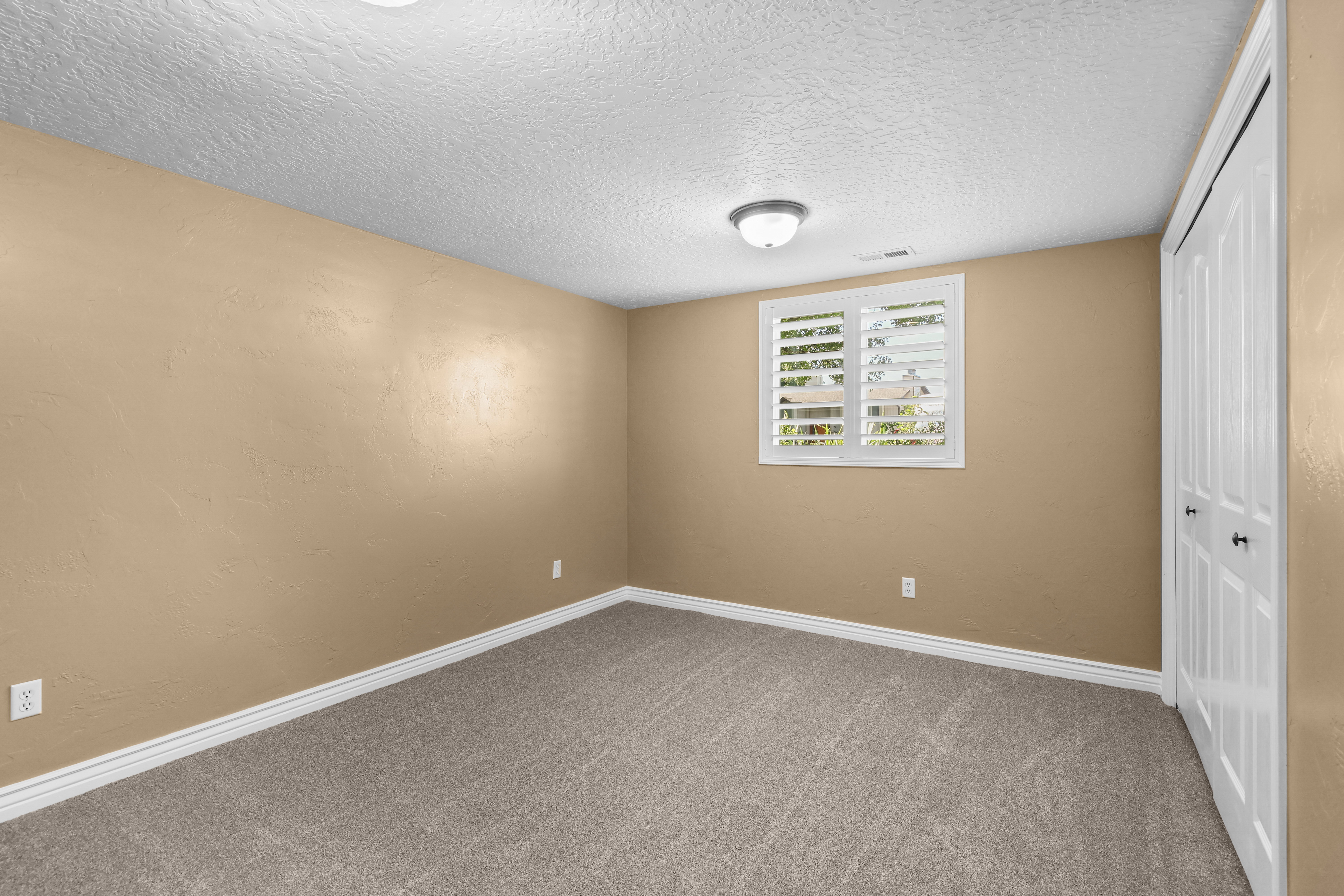 Neutral-toned carpeting in a spacious room featuring a window with white plantation shutters, a surface-mounted ceiling light fixture, and a white bi-fold closet door with black hardware
