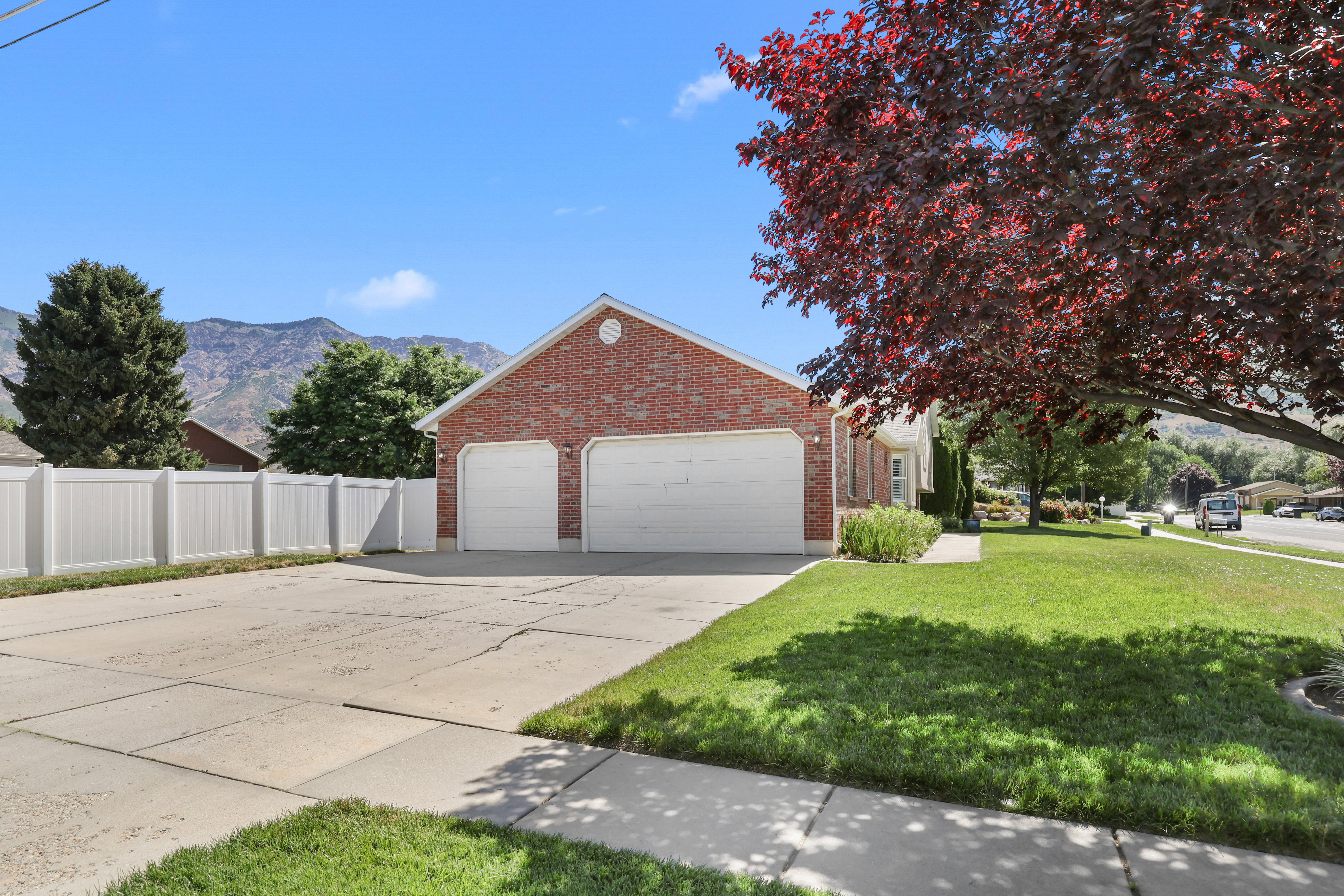Brick exterior featuring a three-car garage with white doors, expansive concrete driveway, manicured lawn, and white vinyl fencing