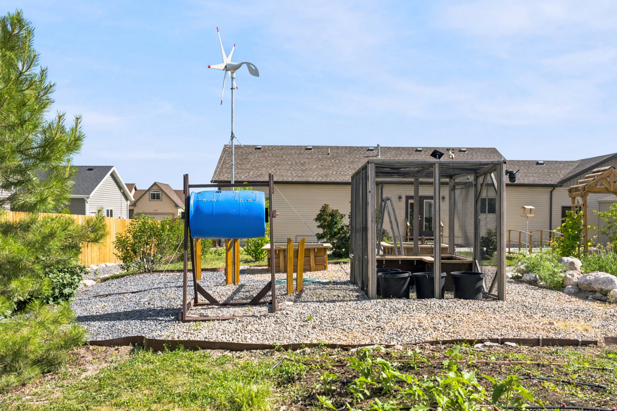Gravel backyard area featuring a compost tumbler, a small wind turbine, and a screened in garden area