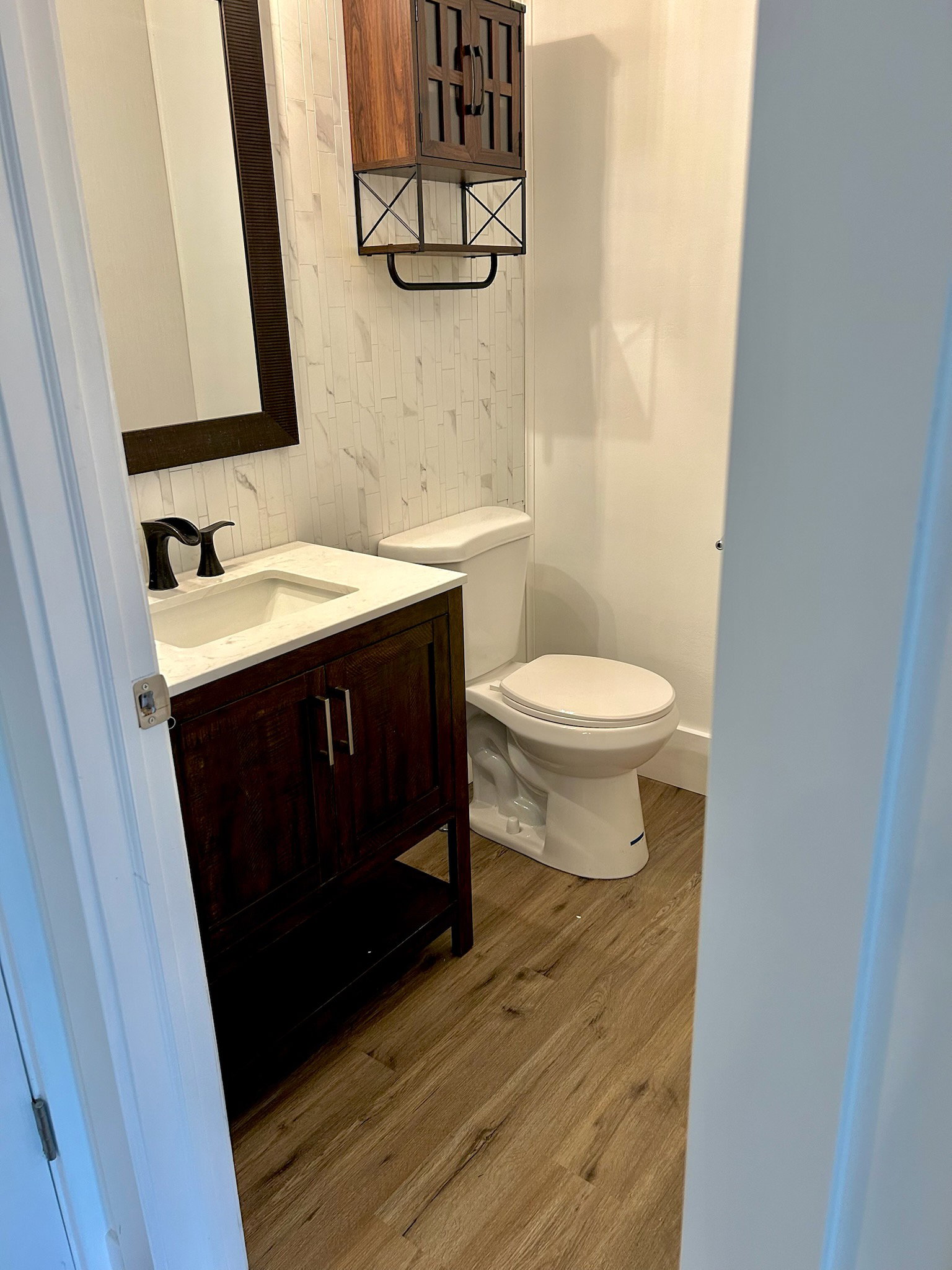 Powder room featuring wood-finish flooring, a dark wood vanity with white countertop and integrated sink, a dark framed mirror, and a wall-mounted wood cabinet with black metal accents