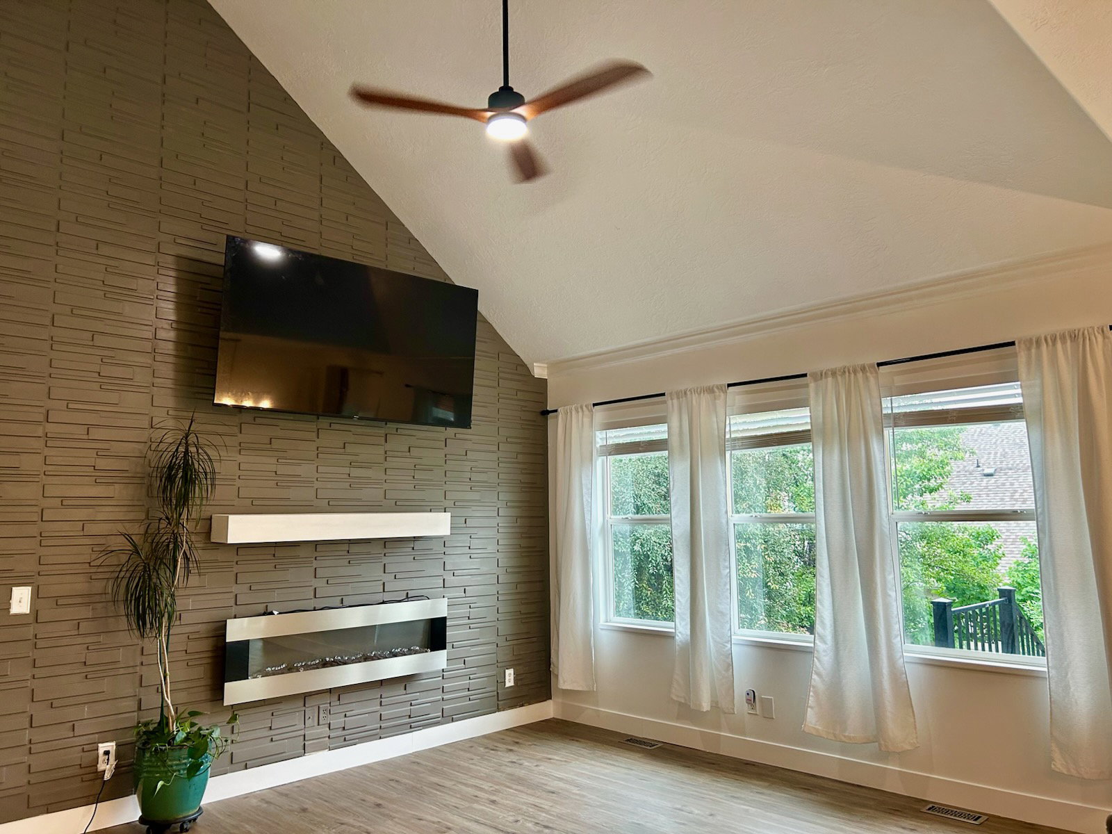 Vaulted ceiling with a contemporary ceiling fan, textured accent wall, linear electric fireplace, three large windows, and wood-finish flooring
