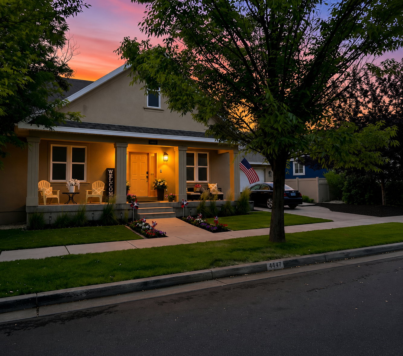 Stucco exterior residence featuring a covered front porch with substantial columns, a prominent front door with sidelights, a paved driveway, and manicured landscaping with planted flower beds