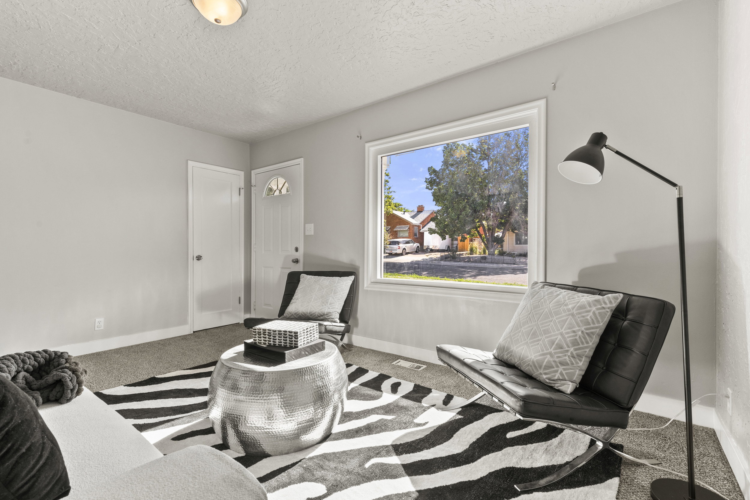 Bright living space featuring a large window, light gray walls, and textured carpeting