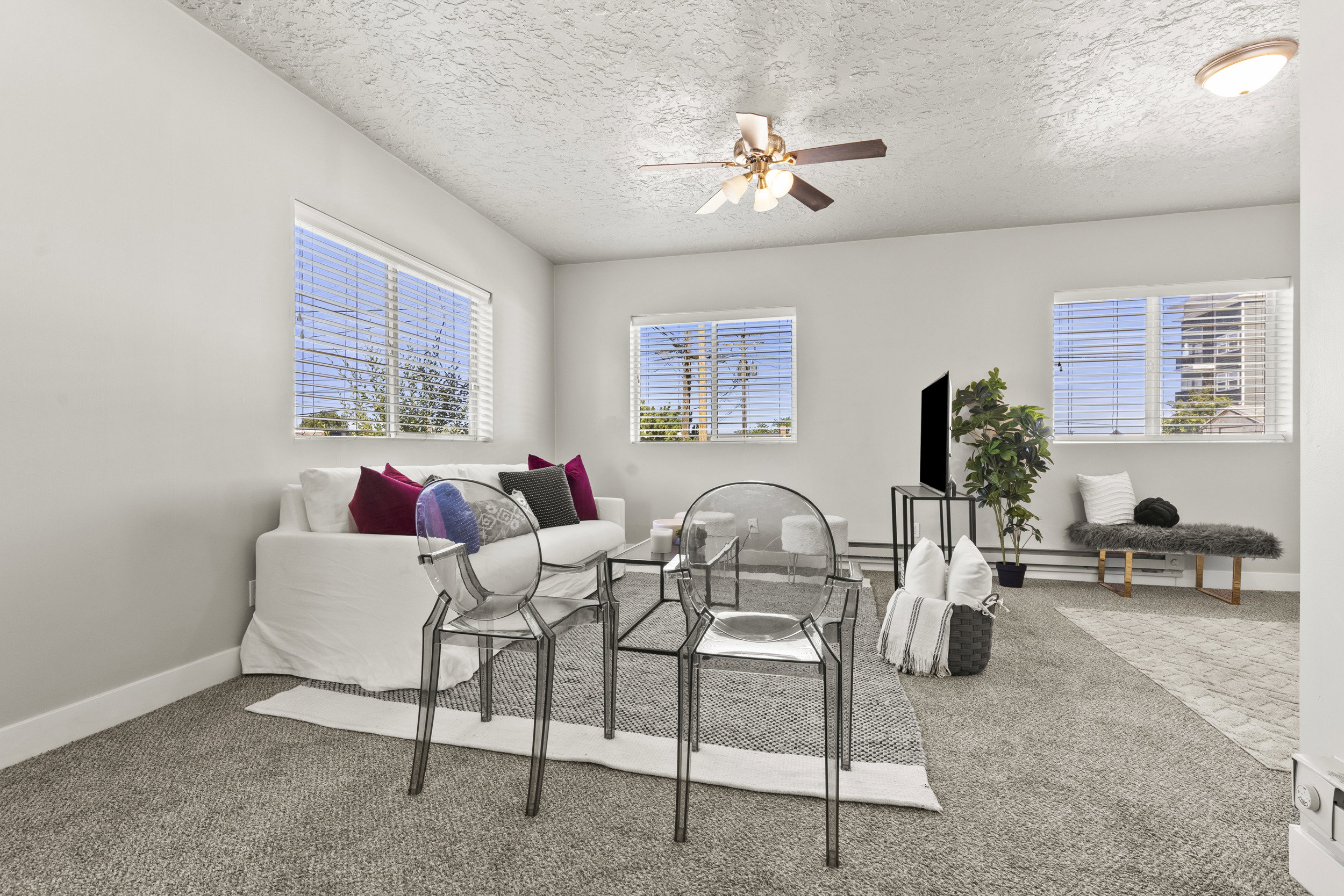 Bright living area featuring light gray carpeting, multiple windows with blinds, a ceiling fan with integrated lighting, and white baseboards