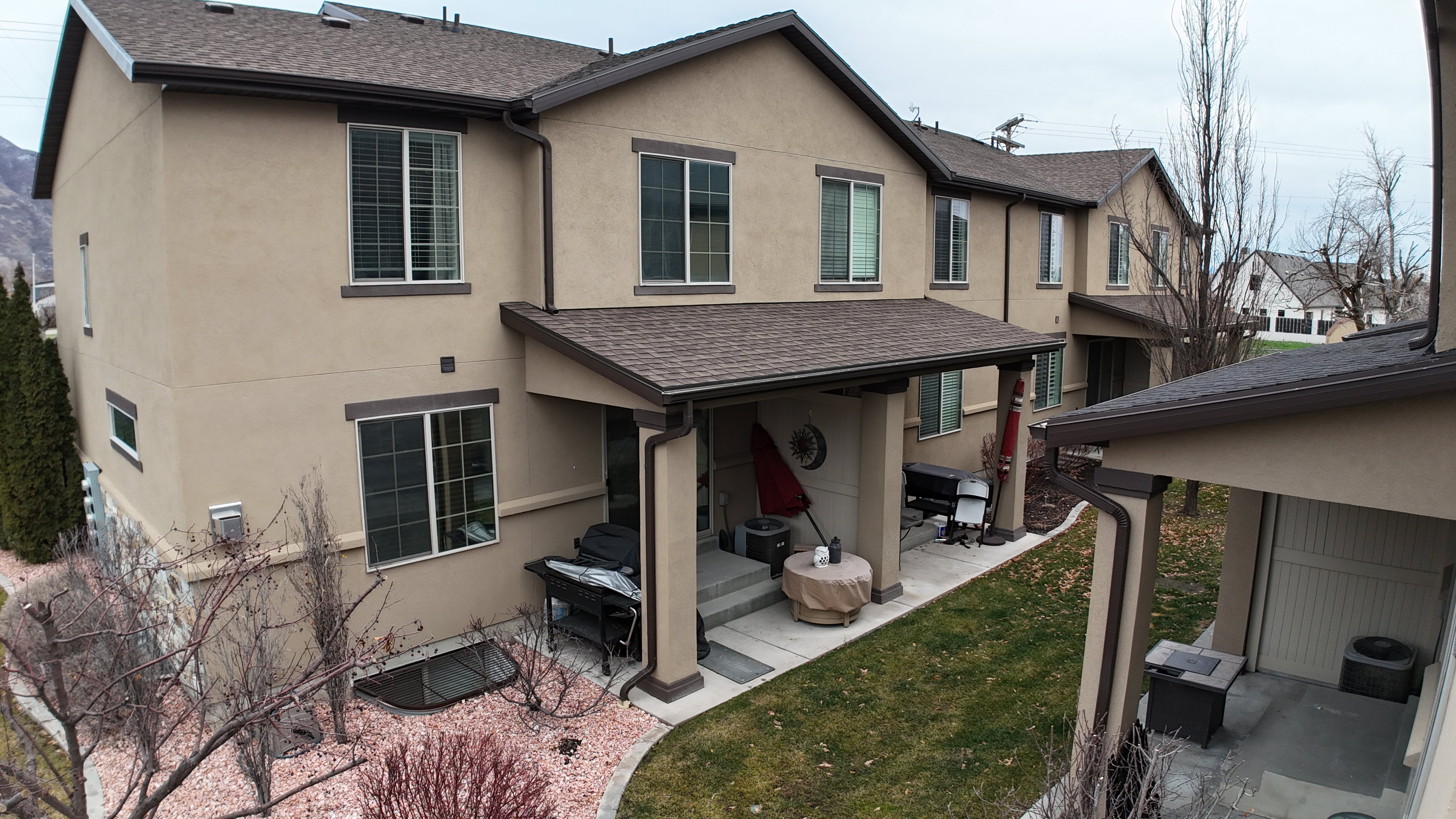 Townhome exterior featuring tan stucco siding with brown trim, multiple windows with blinds, a covered patio, and an adjacent grassy area