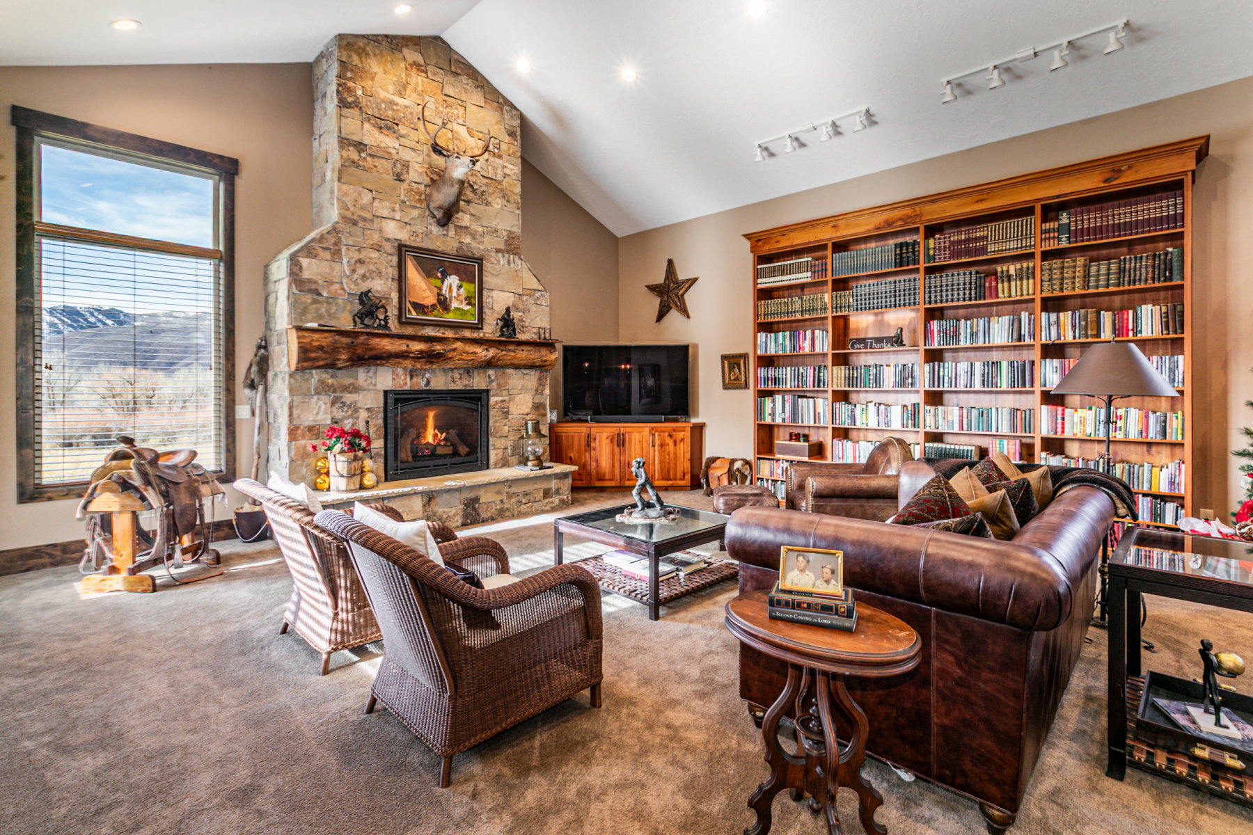 Spacious living area featuring a stone fireplace with a wood mantel, a large window offering exterior views, and extensive built-in wooden bookshelves