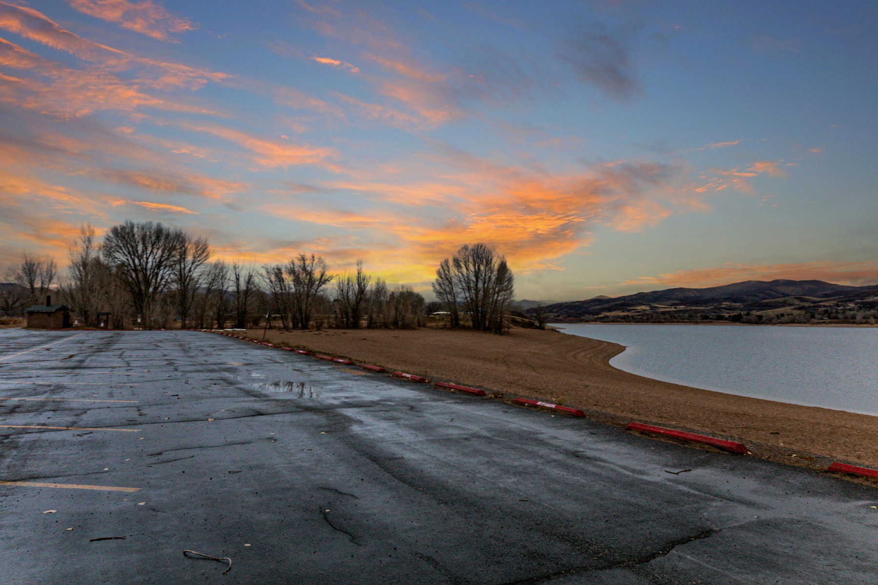 Paved lot with shoreline access to a calm body of water, backed by distant mountains and a tree line