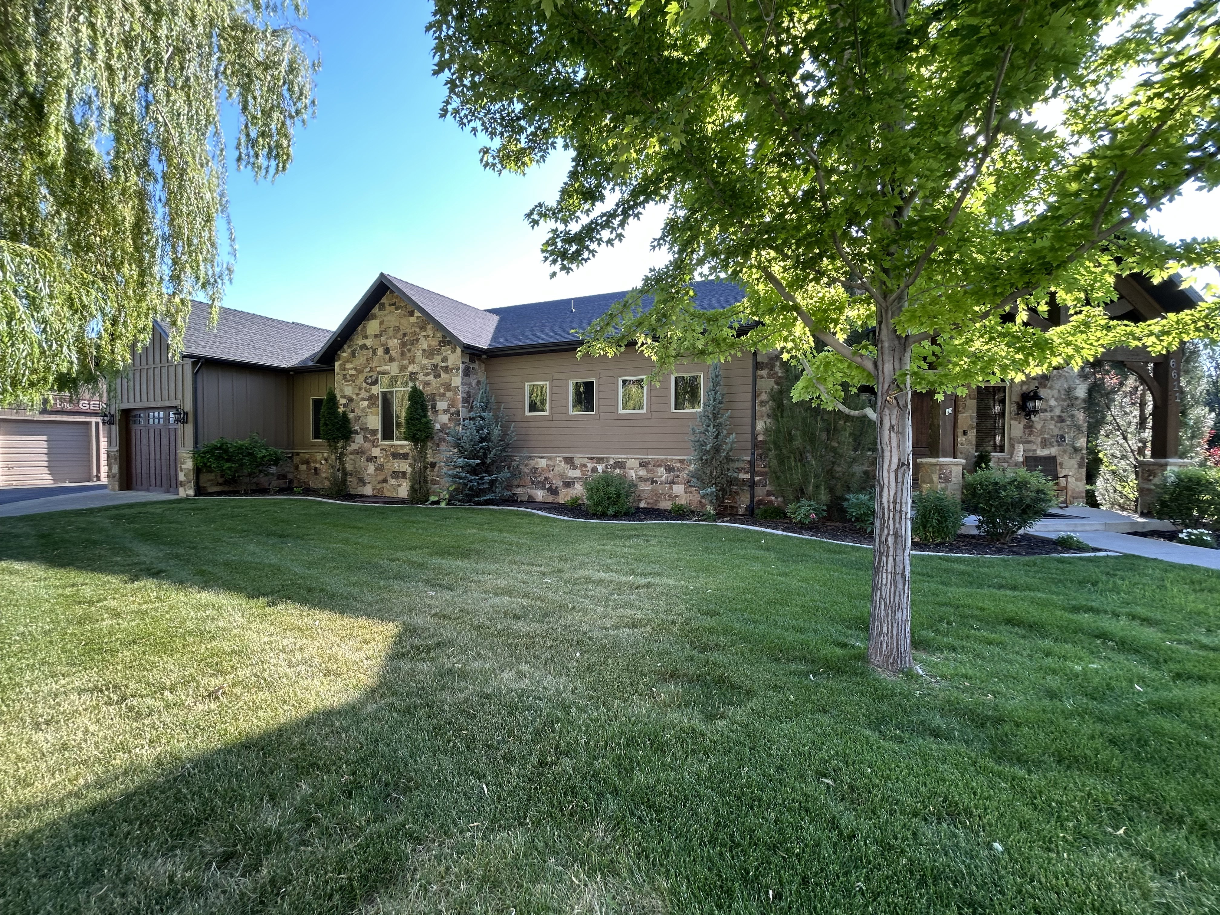 Stone and siding exterior featuring an attached garage with a wood-finish door, a manicured lawn, mature trees, and a paved driveway