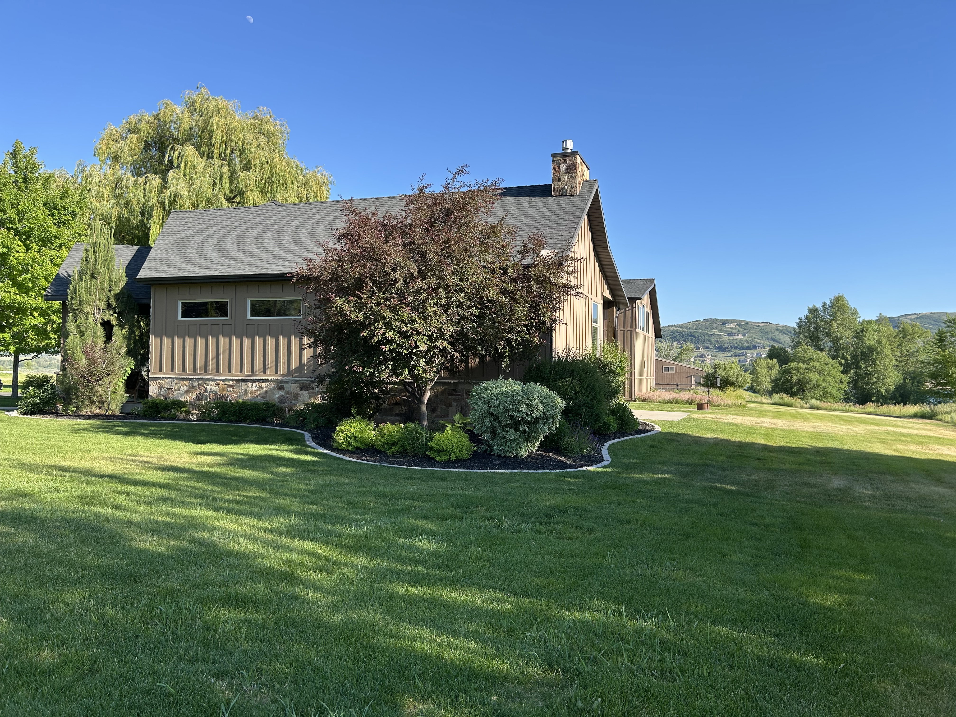 Exterior featuring vertical wood siding, stone facade, a shingled roof, a stone chimney, and a landscaped foundation planting bed