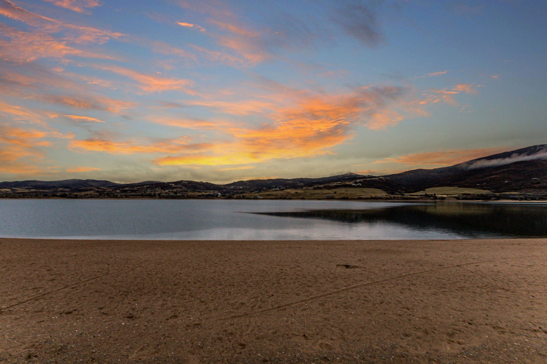 Expansive waterfront with a tranquil lake, sandy shore, and distant rolling hills