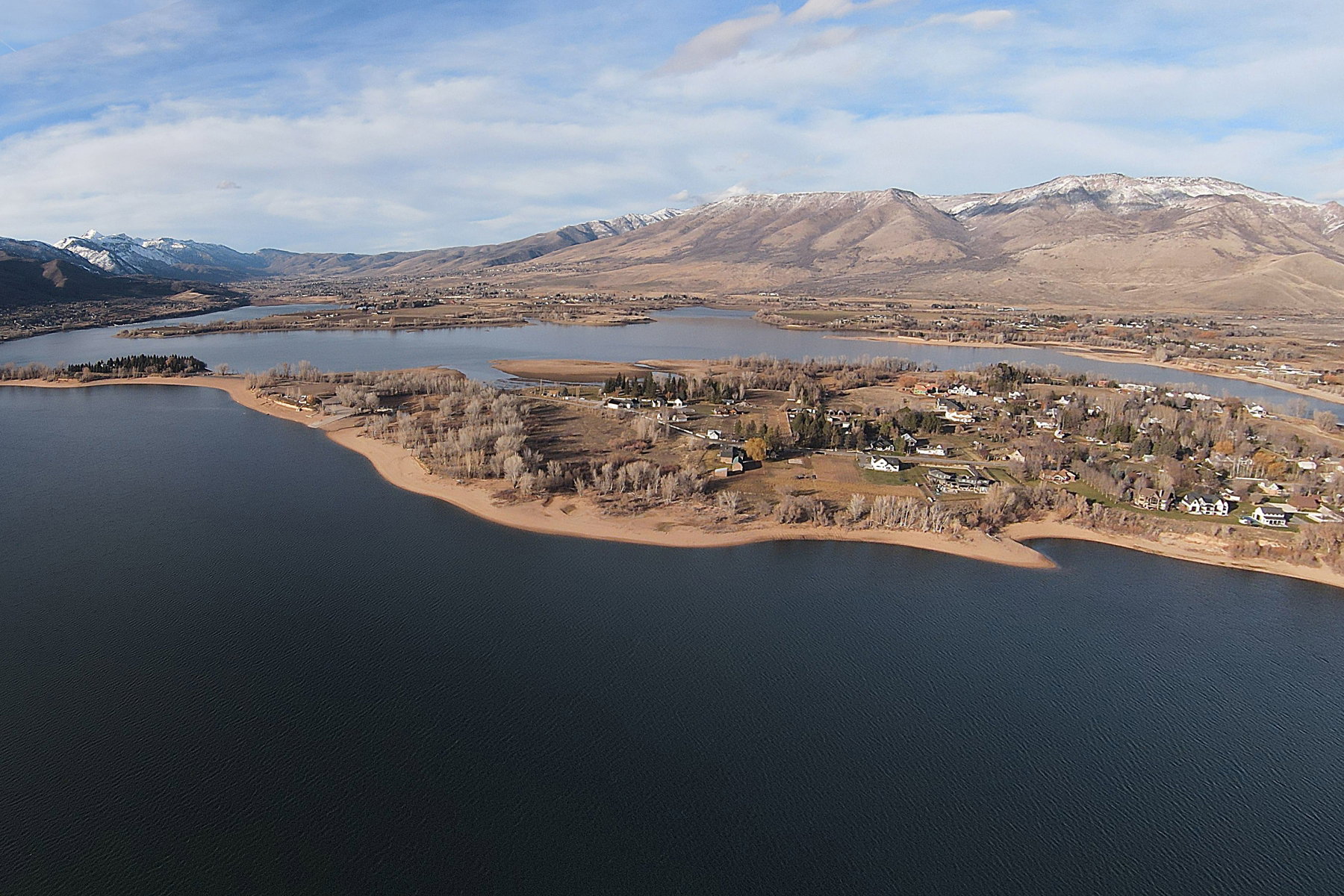 Expansive aerial showcasing a large body of water, a residential area with varied housing styles, surrounding natural landscapes, and distant snow-capped mountains