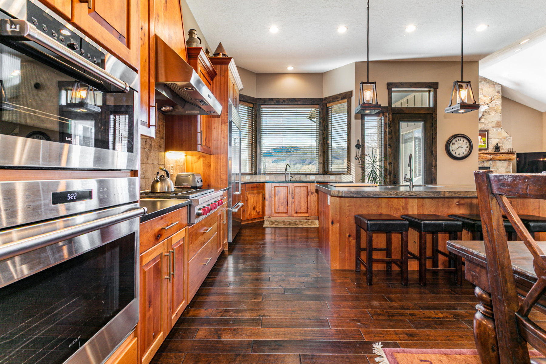 Gourmet kitchen featuring stainless steel double ovens, a built-in range with a professional-grade hood, rich wood cabinetry, wood-finish flooring, and a multi-level island with seating