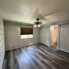 Primary bedroom with view in ensuite Spacious room featuring multi-tone wood-finish flooring, a contemporary ceiling light fixture, a window with a brown roll-down shade, and multiple closet storage options with bi-fold doors