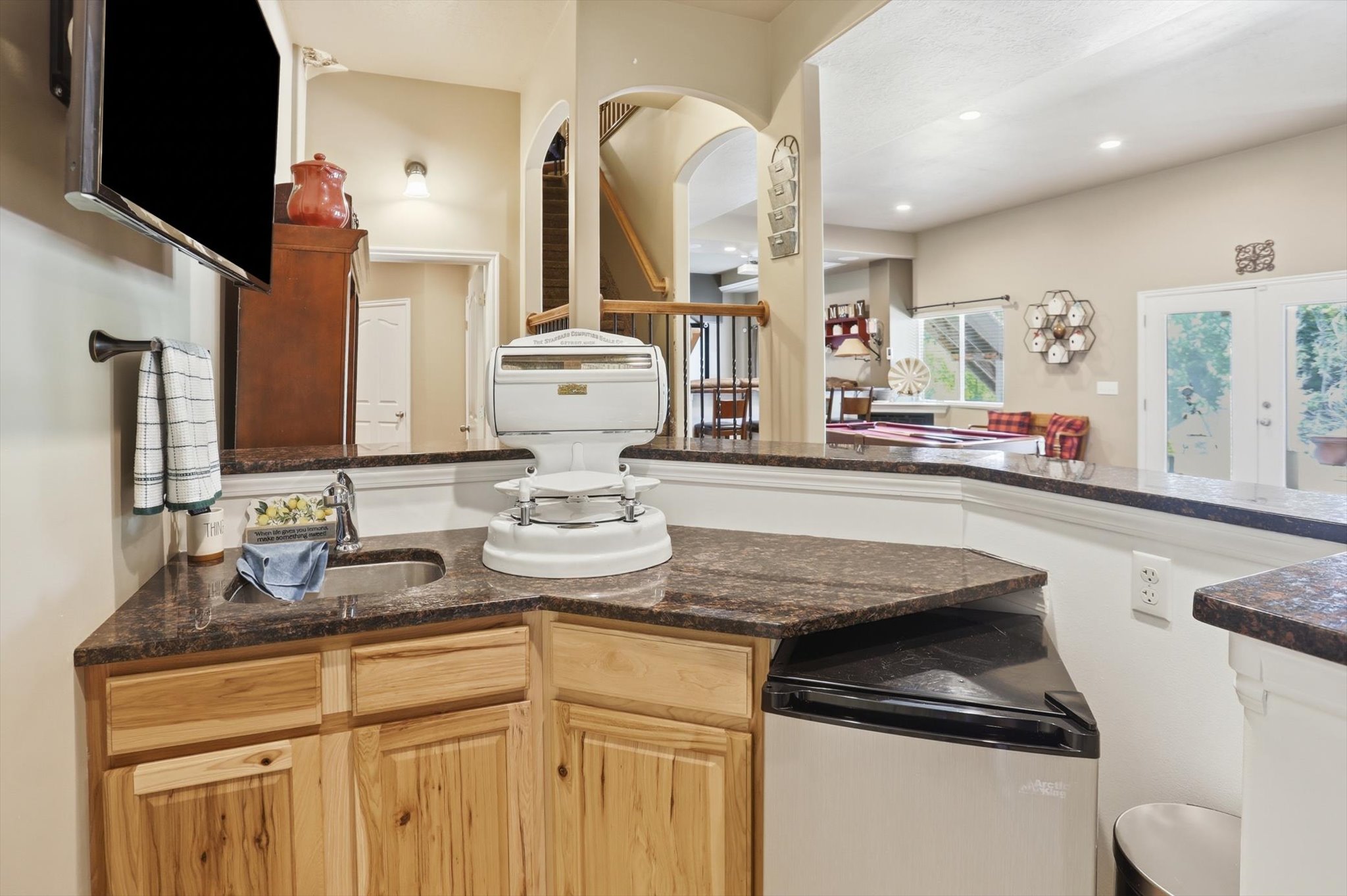 Built-in wet bar featuring a stainless steel sink, dark granite-style countertop, light wood cabinetry, and an under-counter refrigerator