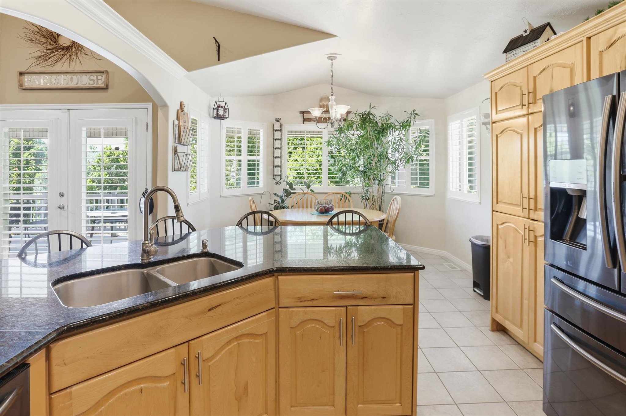 Kitchen island with dual stainless steel sink and dark stone countertop, light wood cabinetry, stainless steel refrigerator, white tile flooring, and dining area with multiple windows