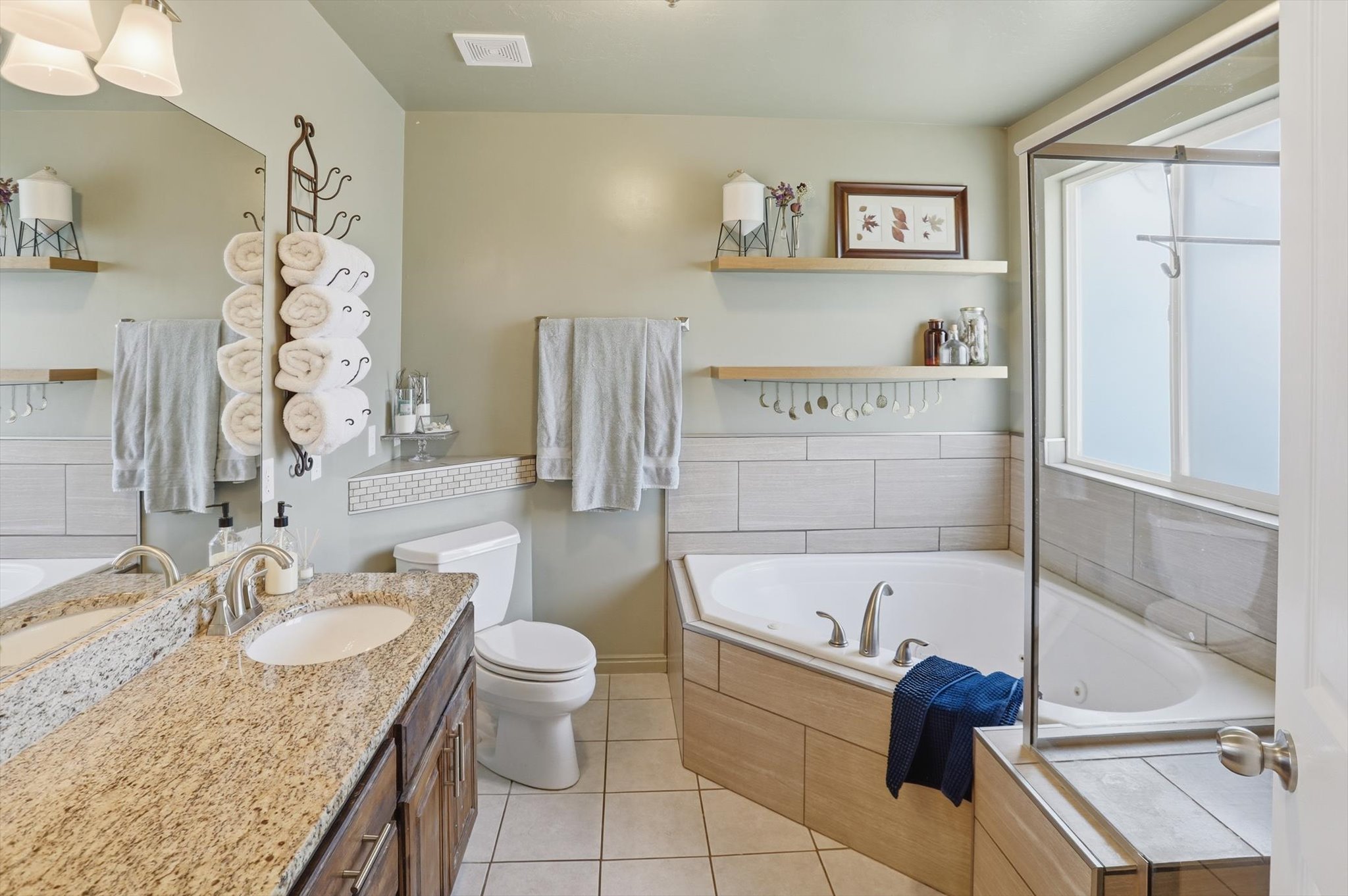 Bathroom featuring a dual-sink vanity with a granite-style countertop, a built-in corner soaking tub with tile surround, a separate glass-enclosed shower, and light-toned tile flooring