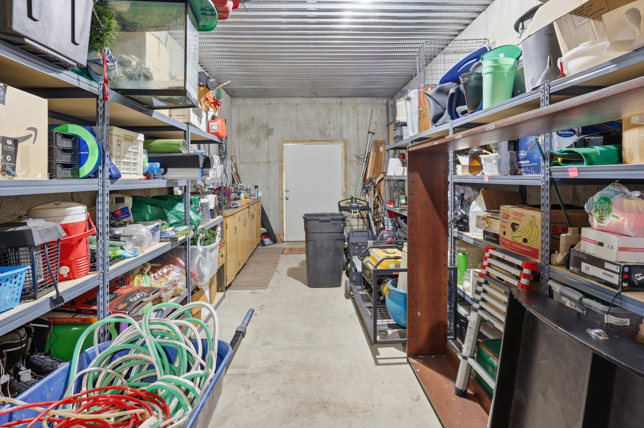 Expansive storage room featuring concrete walls and flooring, a corrugated metal ceiling, and a white door providing access