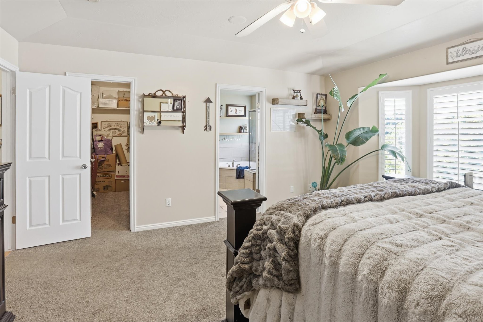 Carpeted room featuring a bay window with plantation shutters, a ceiling fan with integrated lighting, and an open doorway revealing a bathroom with a tiled bathtub surround