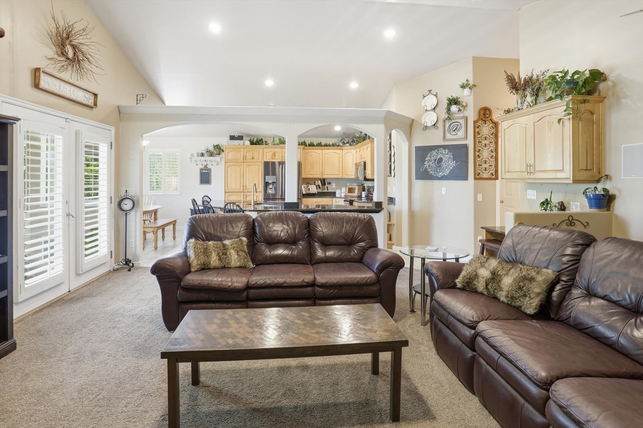 Vaulted ceilings and recessed lighting illuminate this carpeted living space, featuring white louvered patio doors and arched doorways leading to the kitchen