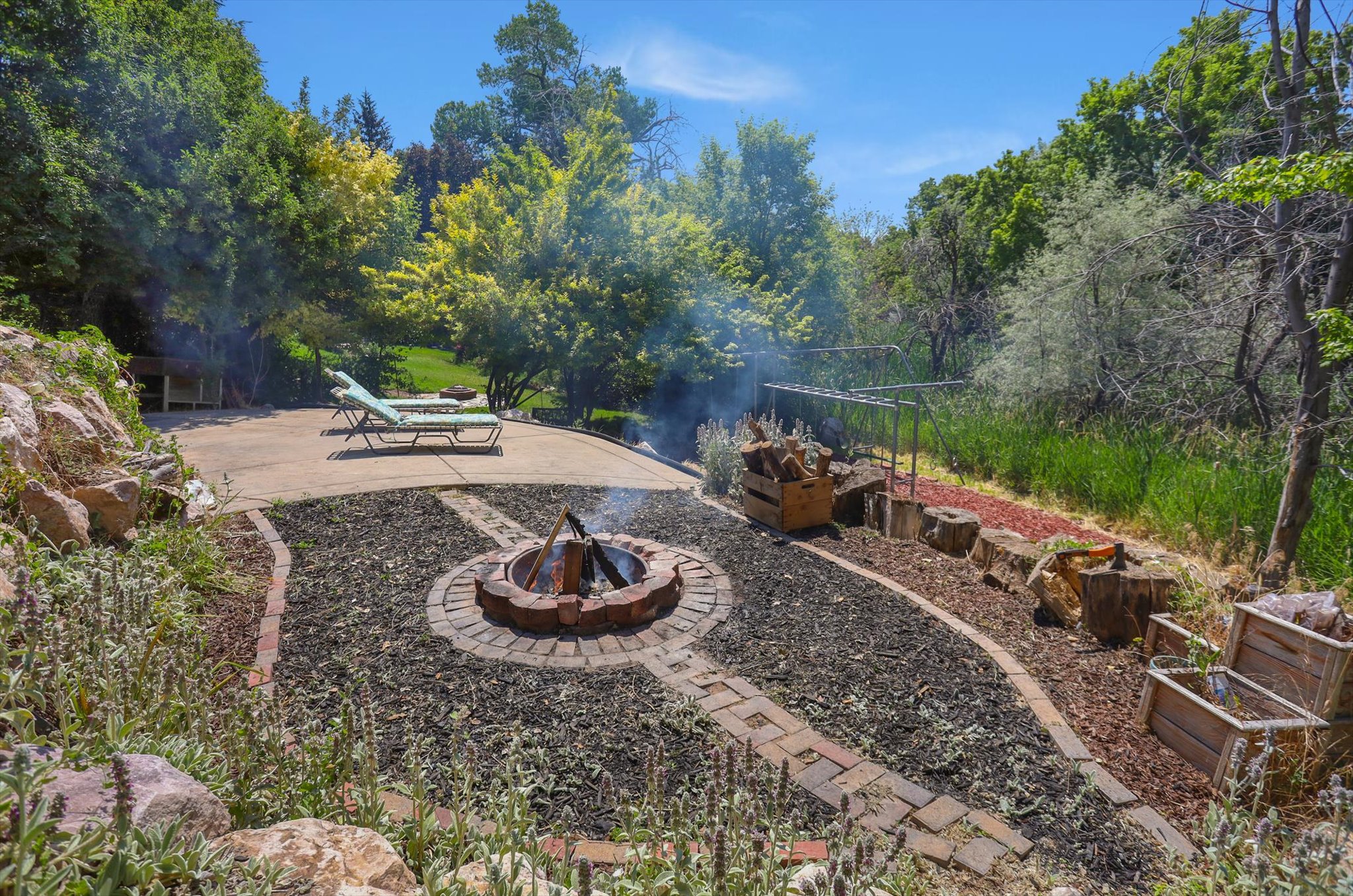 Outdoor patio space featuring a circular brick fire pit, a paved lounging area, and extensive natural landscaping