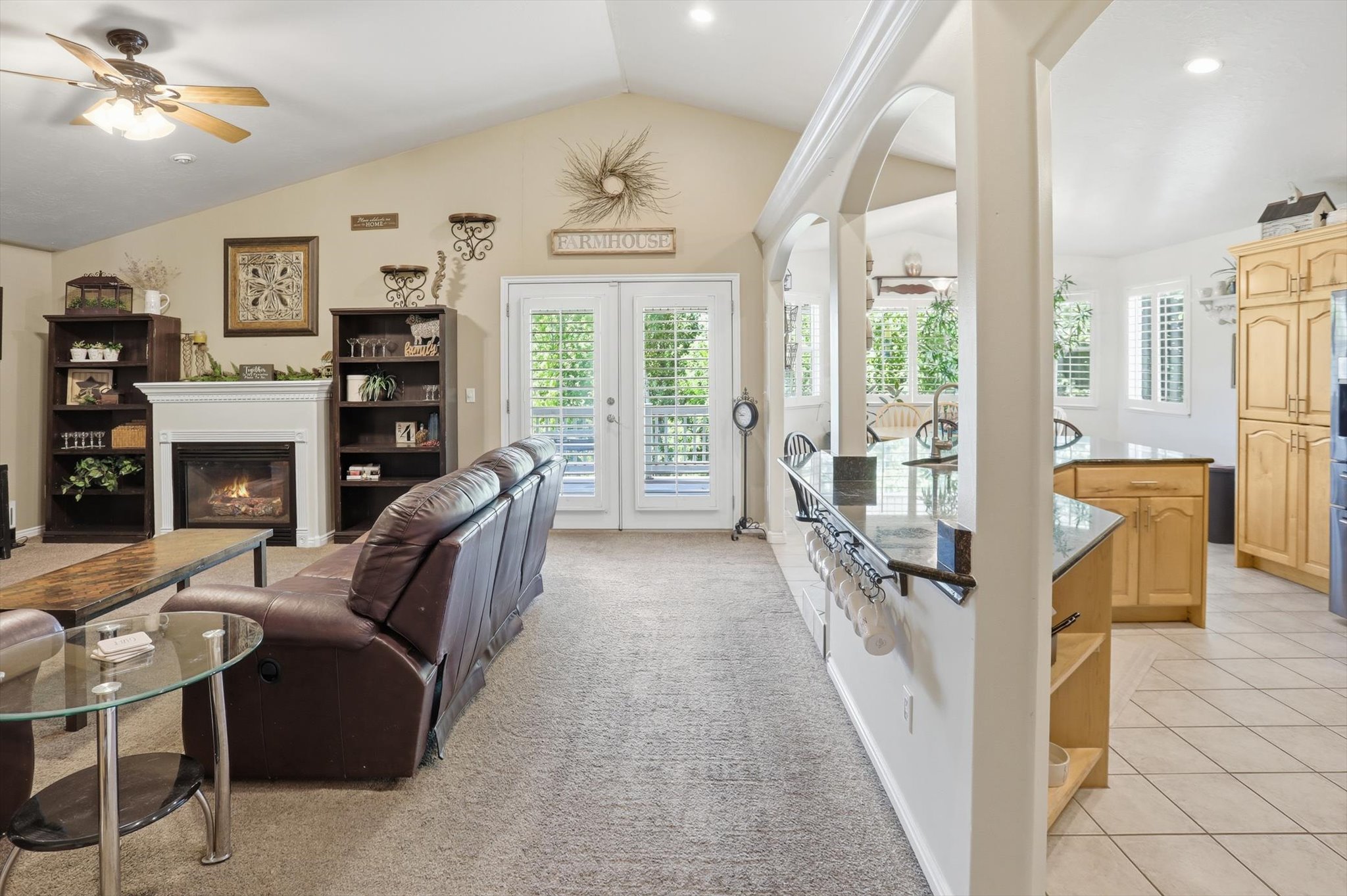 Spacious living area featuring a vaulted ceiling with a ceiling fan, a fireplace with a white mantel, and double glass-paneled doors
