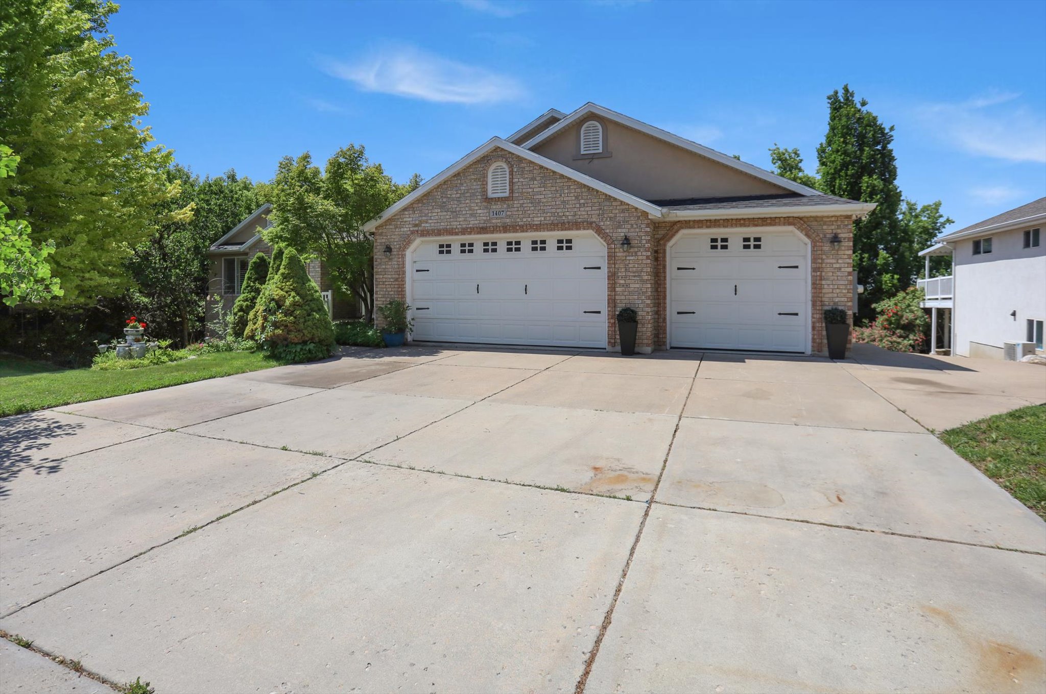 Brick and stucco exterior featuring two overhead garage doors, a concrete driveway, and surrounding mature trees