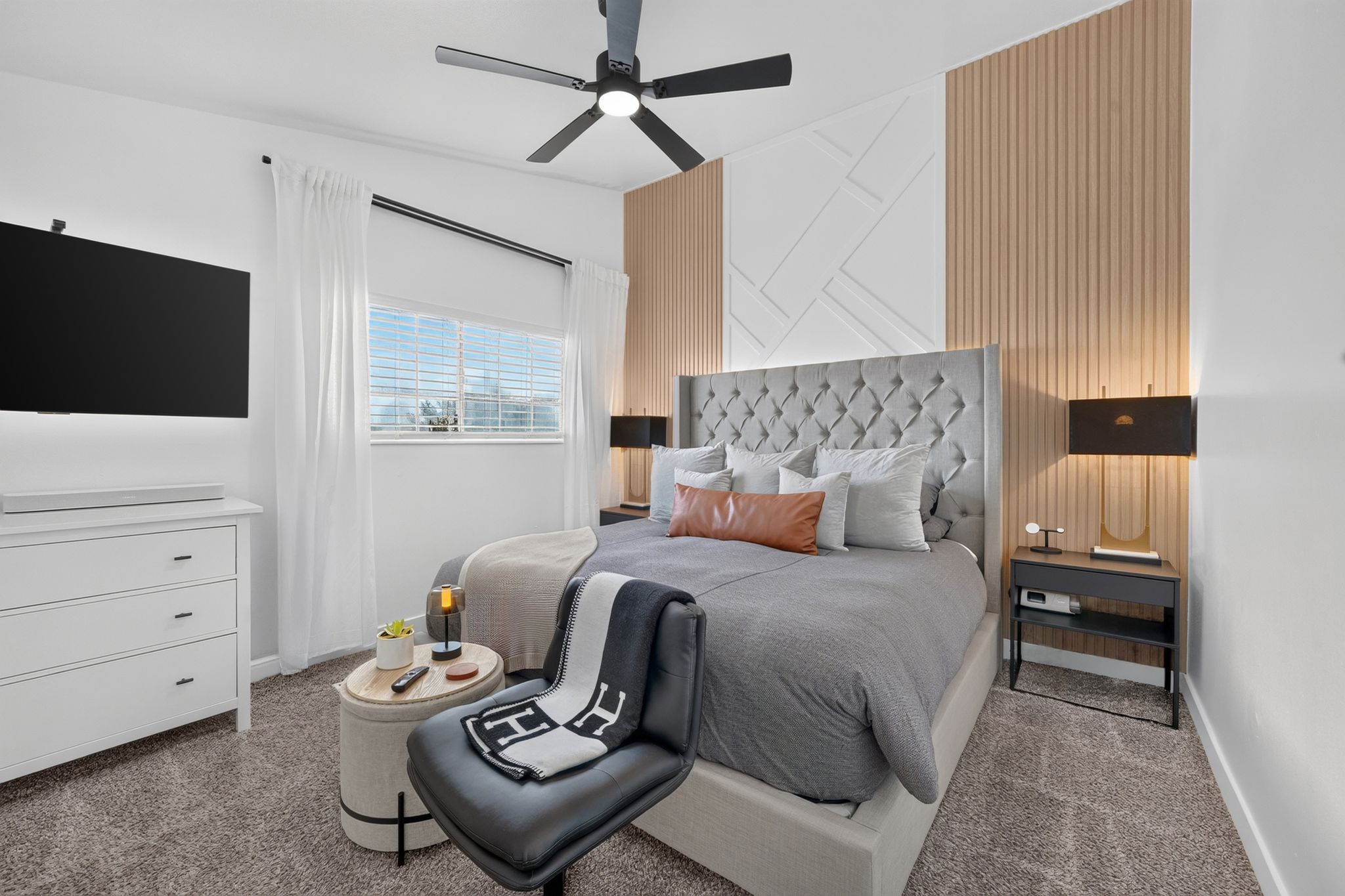 Accent wall with geometric and vertical paneling, plush carpet flooring, a ceiling fan with integrated lighting, a window with blinds and drapes, and a built-in wall-mounted television