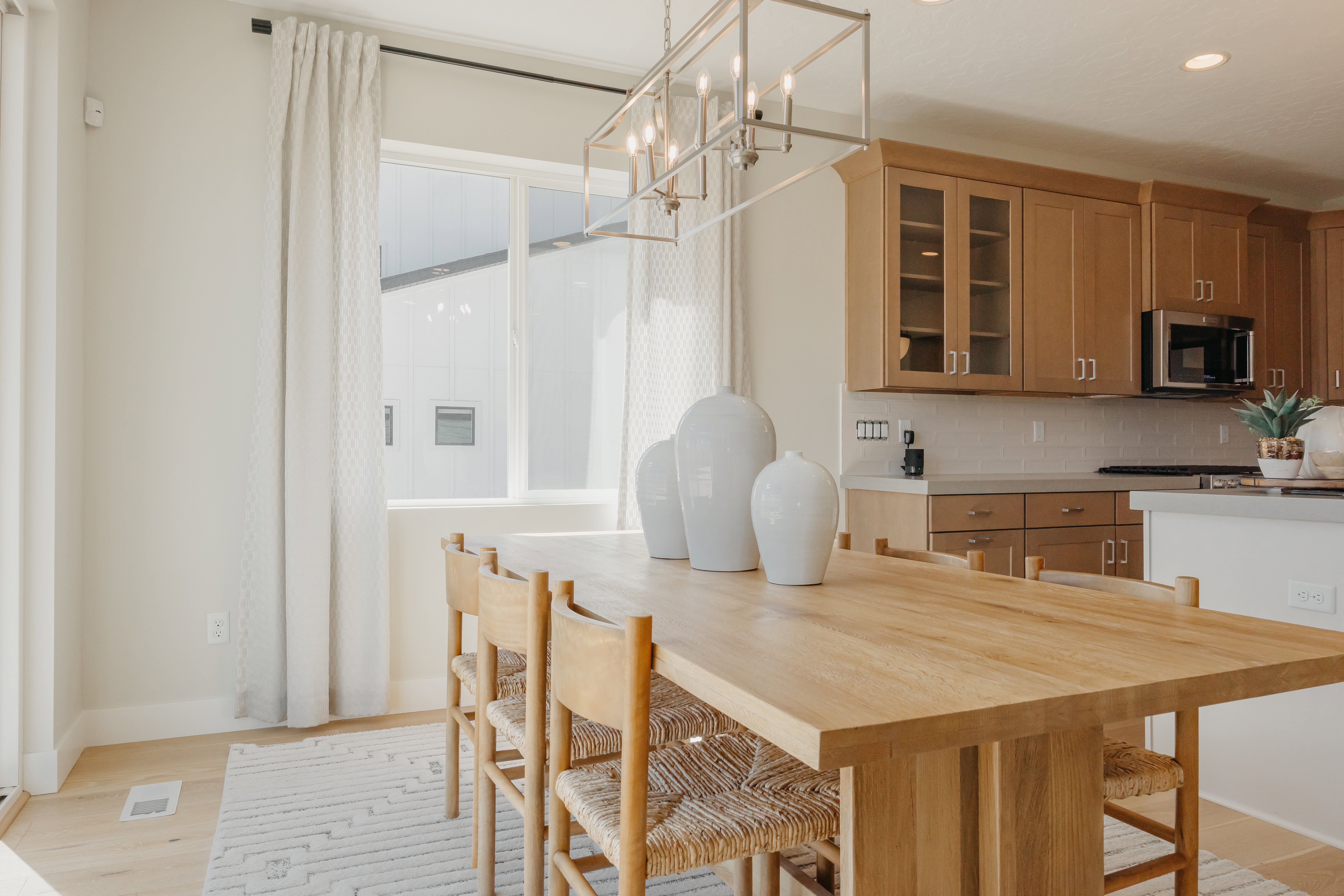 Open-concept dining area featuring light wood-finish flooring, a contemporary linear chandelier, and a bright window with full-length drapes