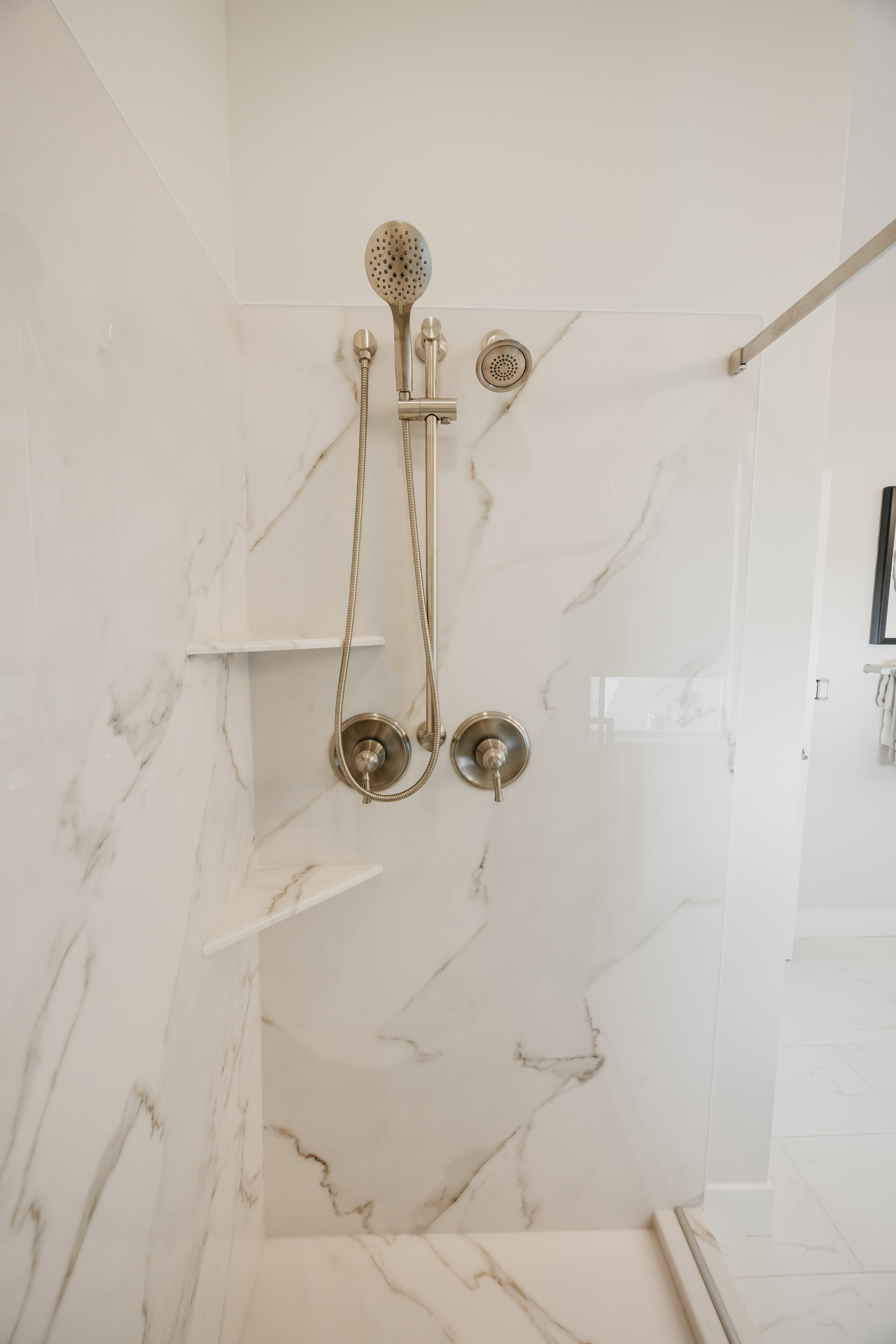 Shower featuring large format marble-look tile, brushed nickel fixtures, a handheld showerhead, and integrated corner shelving