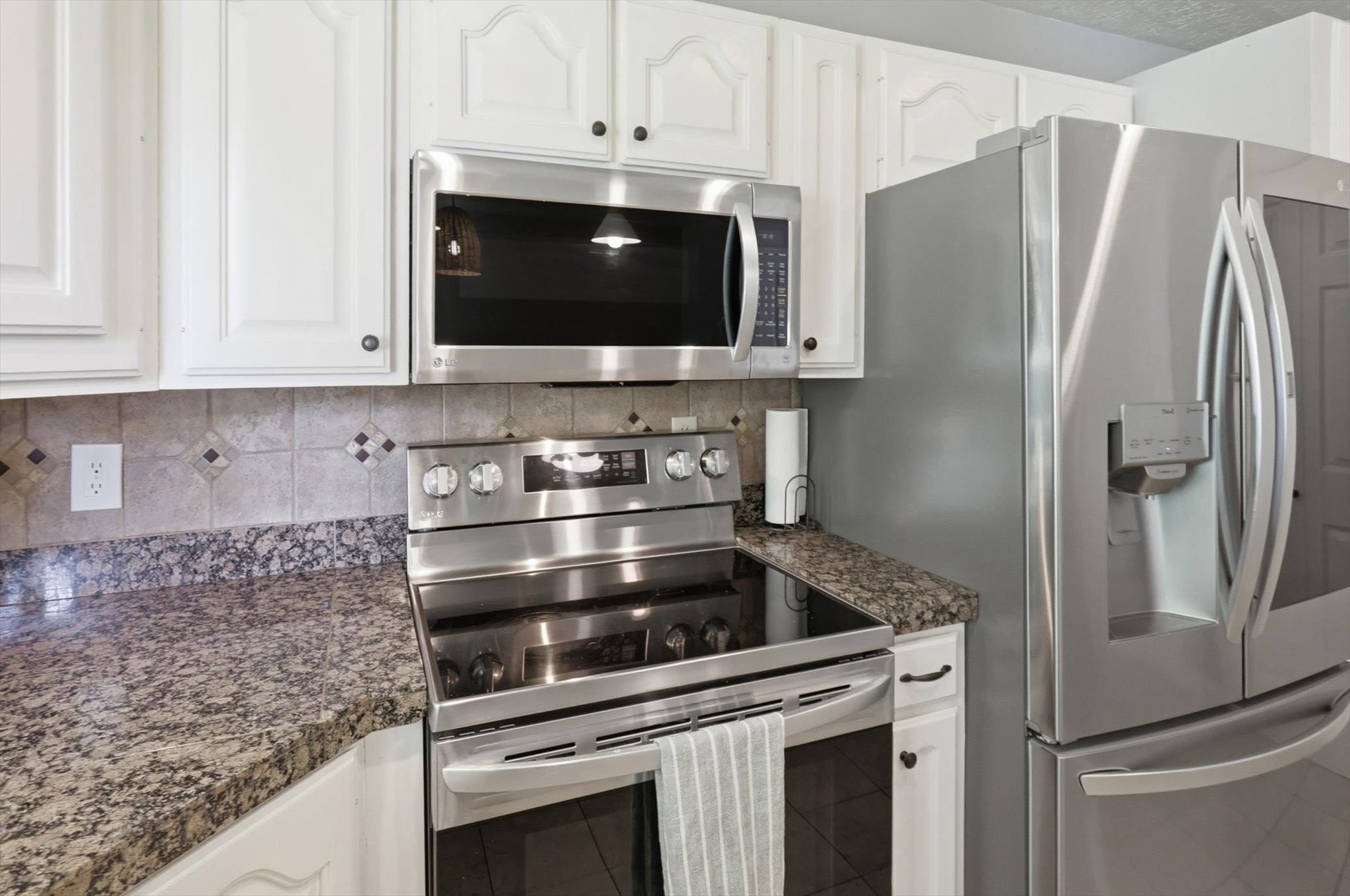 Kitchen featuring white cabinetry, granite-look countertops, tiled backsplash, and stainless steel appliances including a refrigerator, range, and microwave