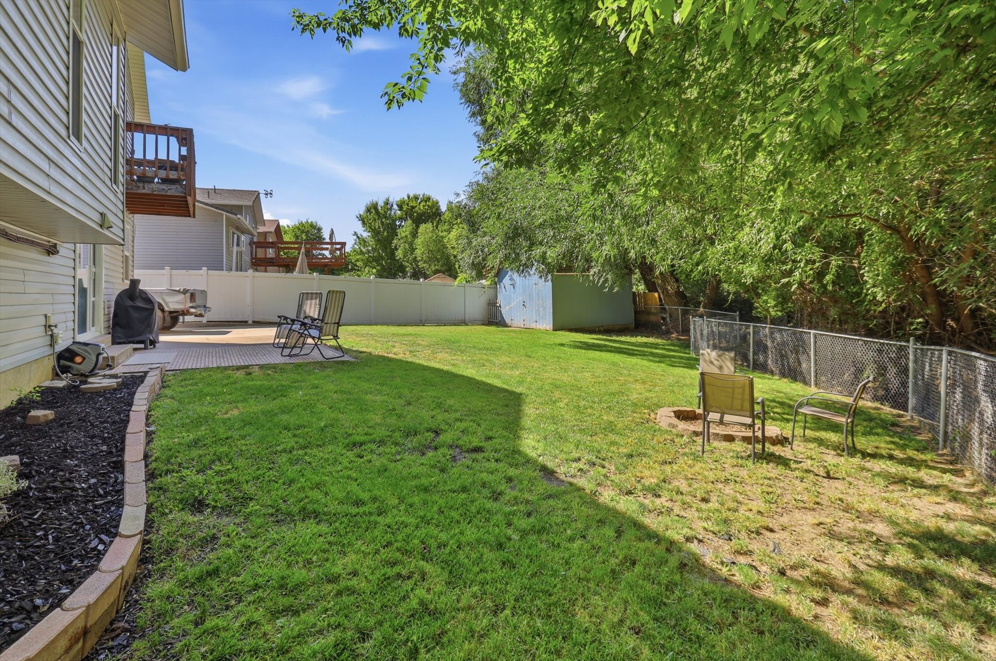 Expansive backyard featuring a stone paver patio, a raised wood deck, a decorative landscape border, and a chain-link fence