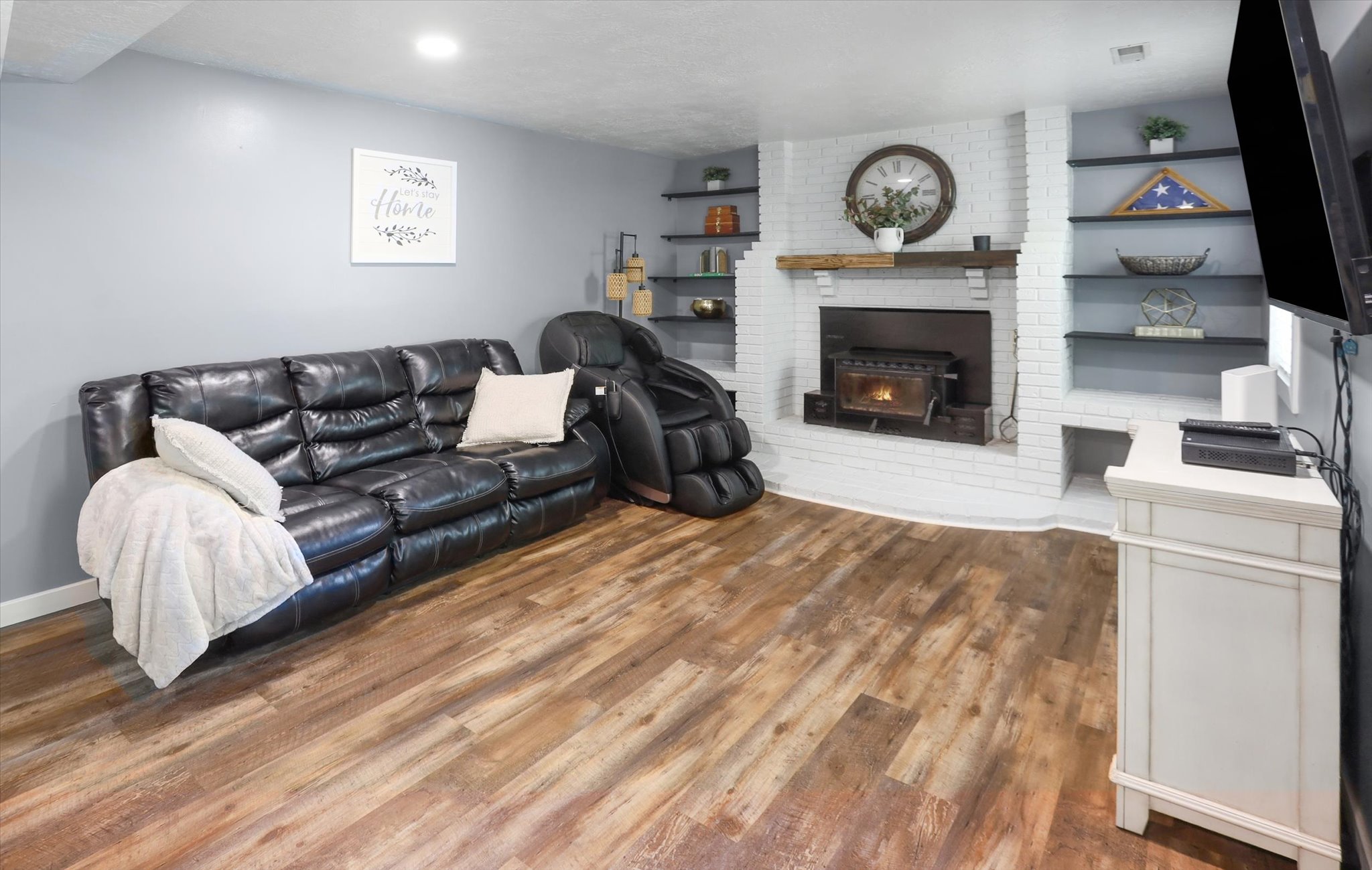 Living area featuring durable wood-finish flooring, a white-painted brick fireplace with a wood-burning insert, a rustic wood mantel, and built-in shelving