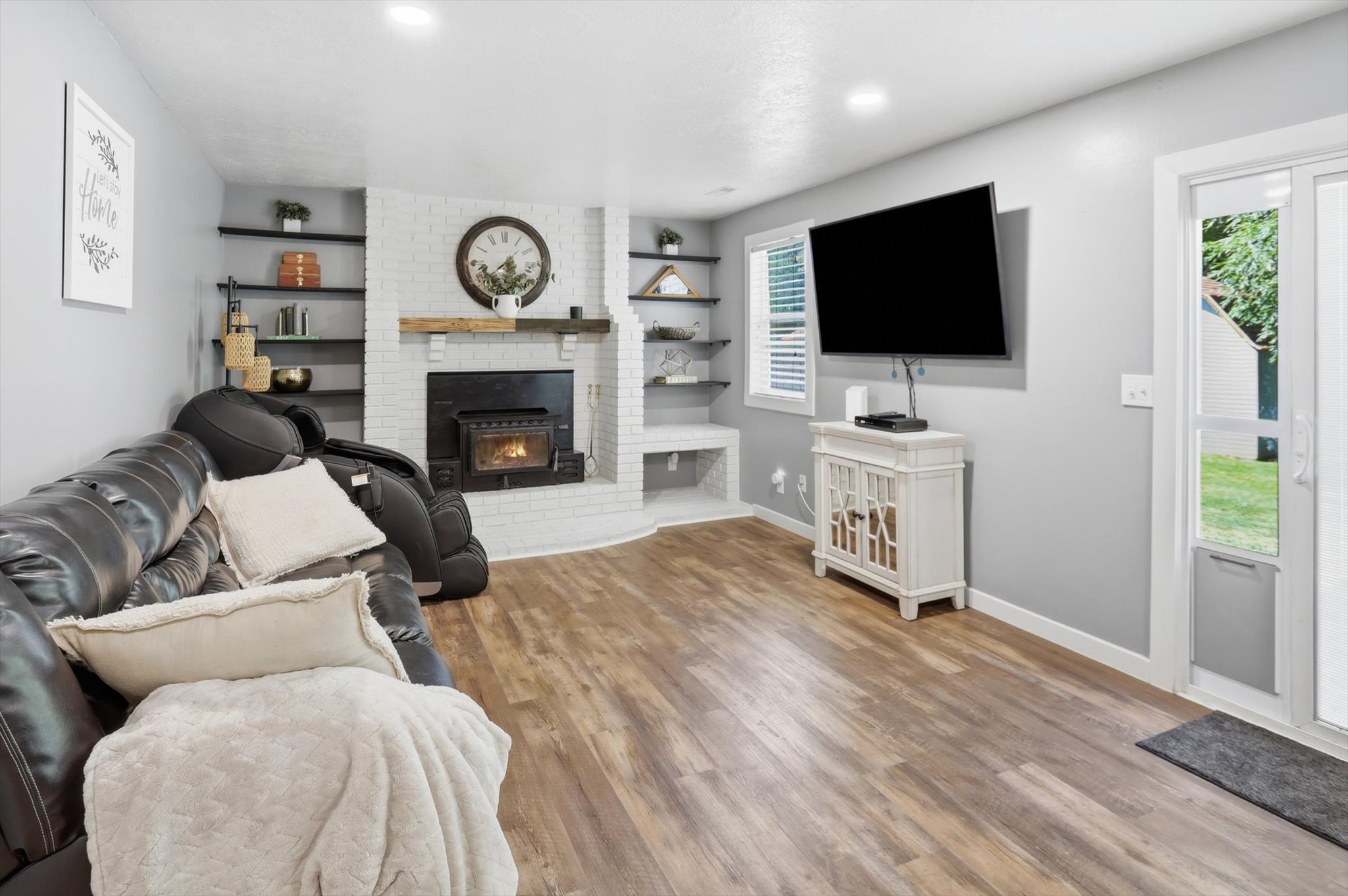 Living area featuring wood-finish flooring, a white brick fireplace with a wood mantle, built-in shelving, recessed lighting, and a glass-paneled exterior door