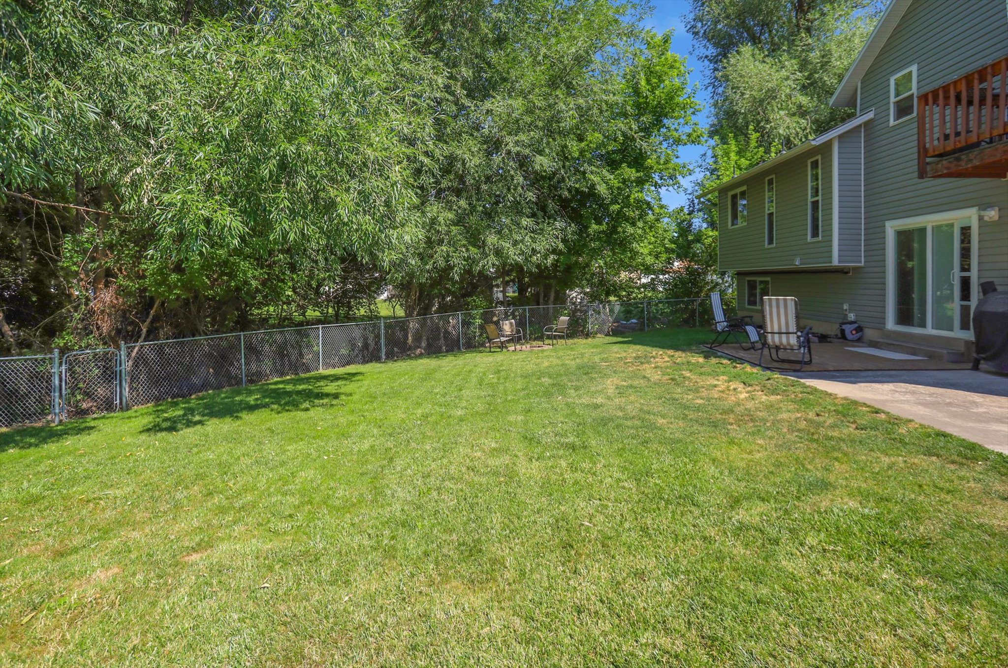 Expansive backyard featuring a green lawn, mature trees, chain-link fencing, and a concrete patio with sliding glass door access to the residence
